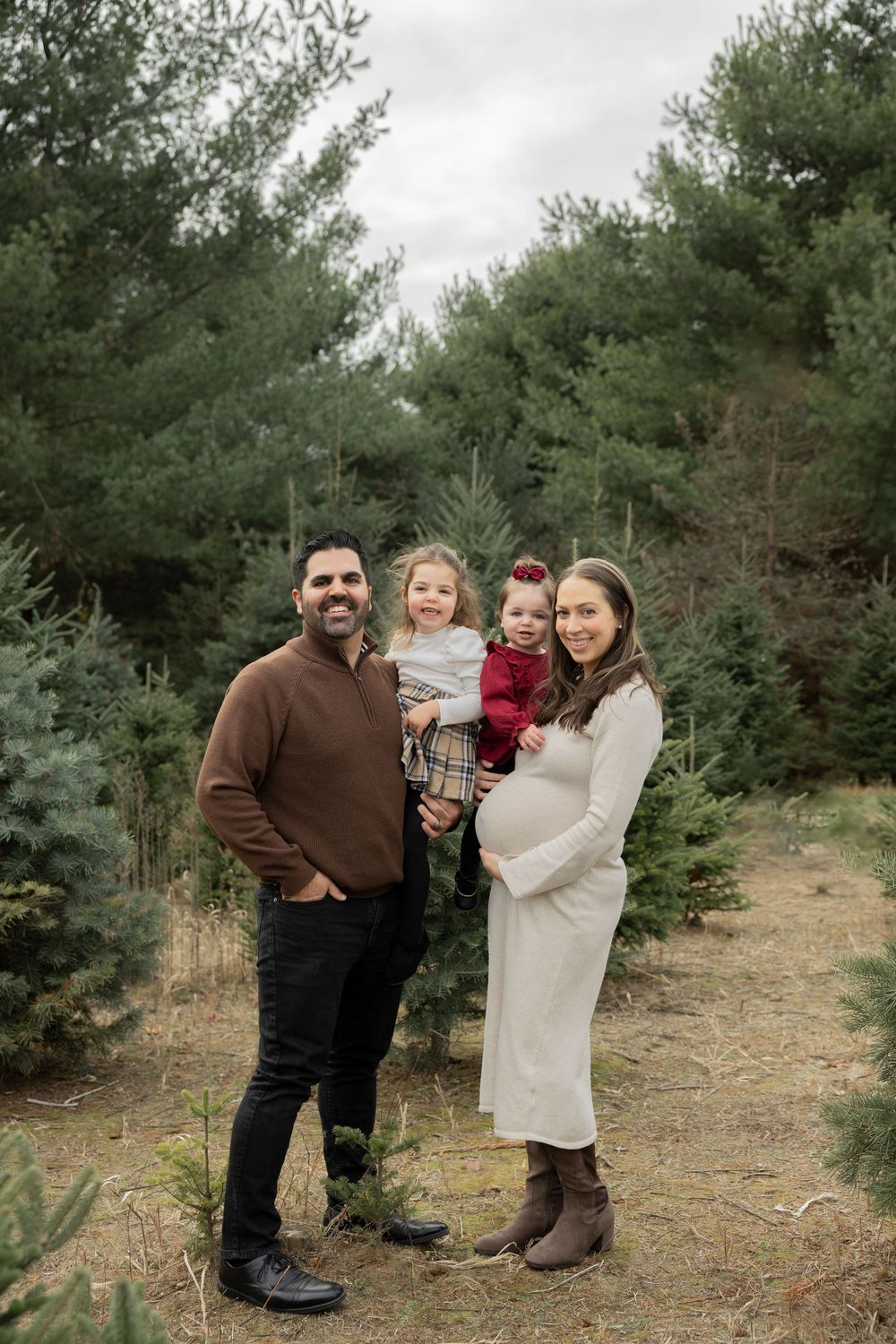 A family poses together at a Christmas tree farm surrounded by evergreen trees on a winter afternoon.