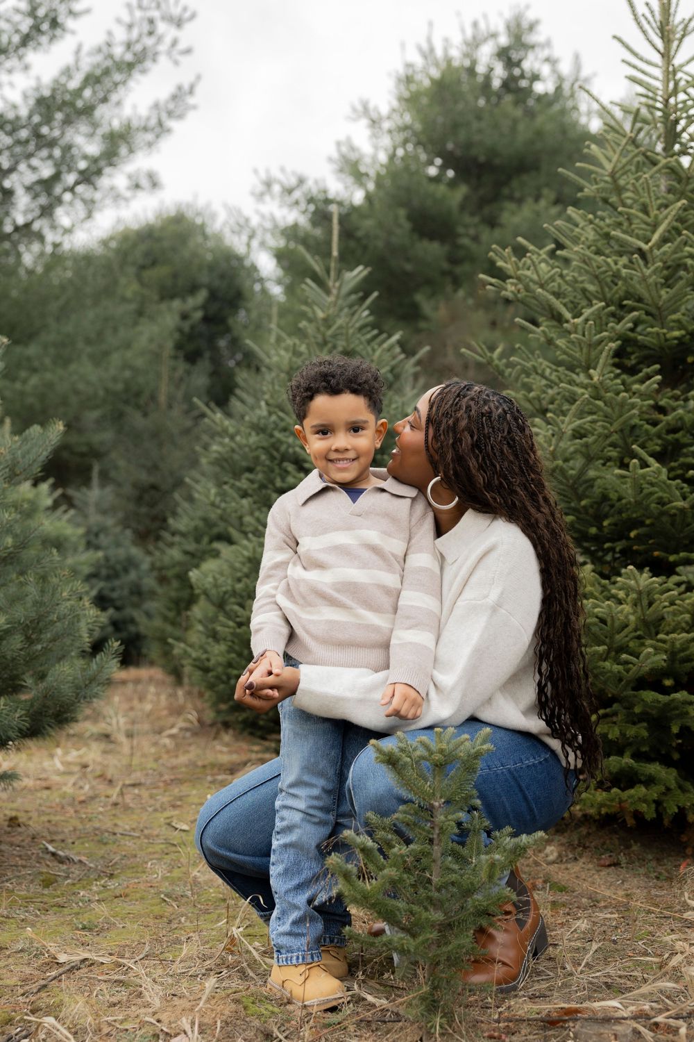 A mother and child share a tender moment while sitting among evergreen trees at a Christmas tree farm.