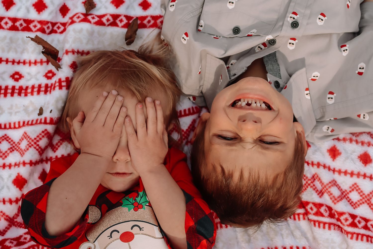 Two children wearing festive holiday pajamas lay on red and white patterned blanket playing peekaboo.