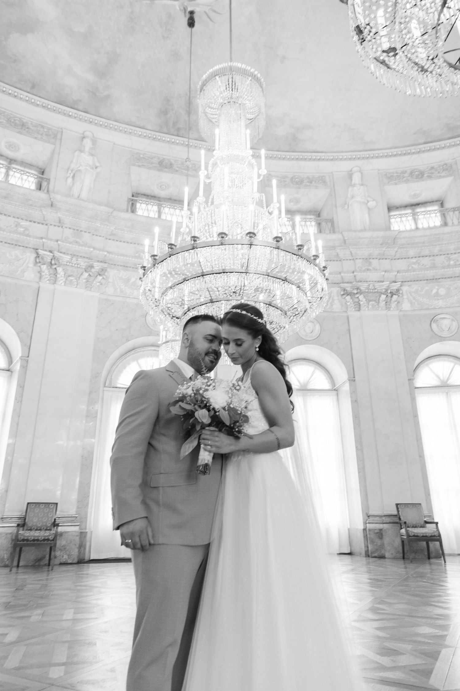 A romantic moment between newlyweds in an elegant white ballroom with ornate chandeliers and classical architecture.