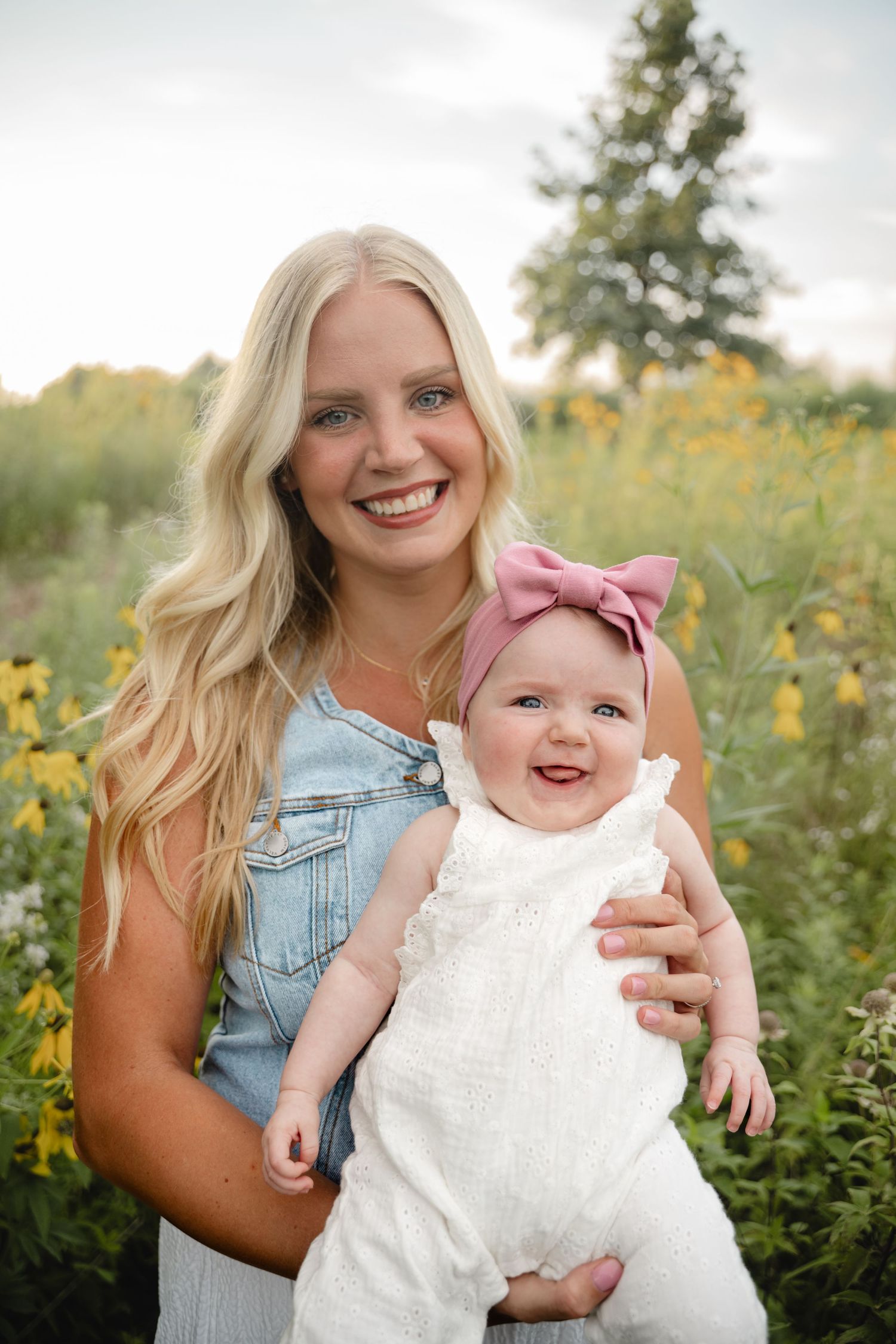 Mother and baby in matching white outfits pose together among yellow wildflowers.