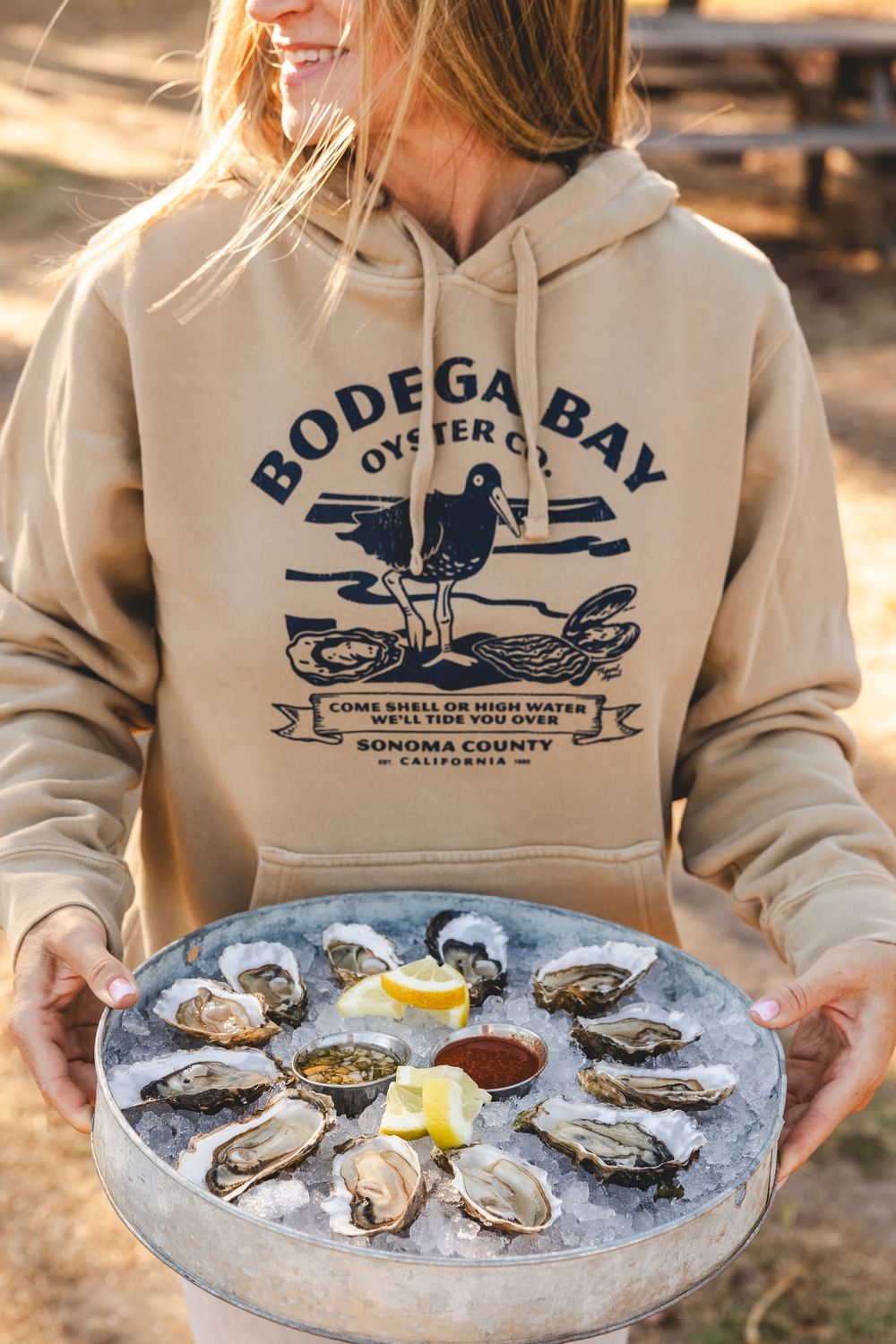 A plate of fresh oysters on ice is served alongside a beige sweatshirt featuring a Bodega Bay nautical design.