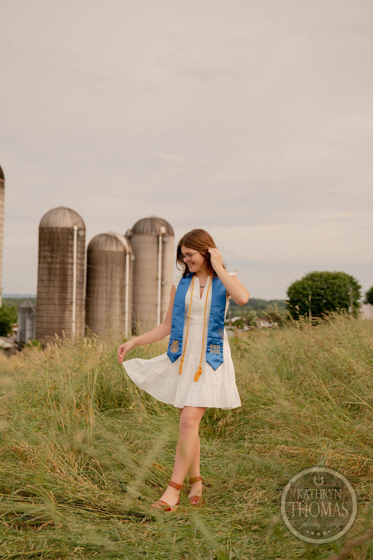 A white sundress and blue jean vest outfit styled against rustic farm silos and tall grass in a countryside setting.
