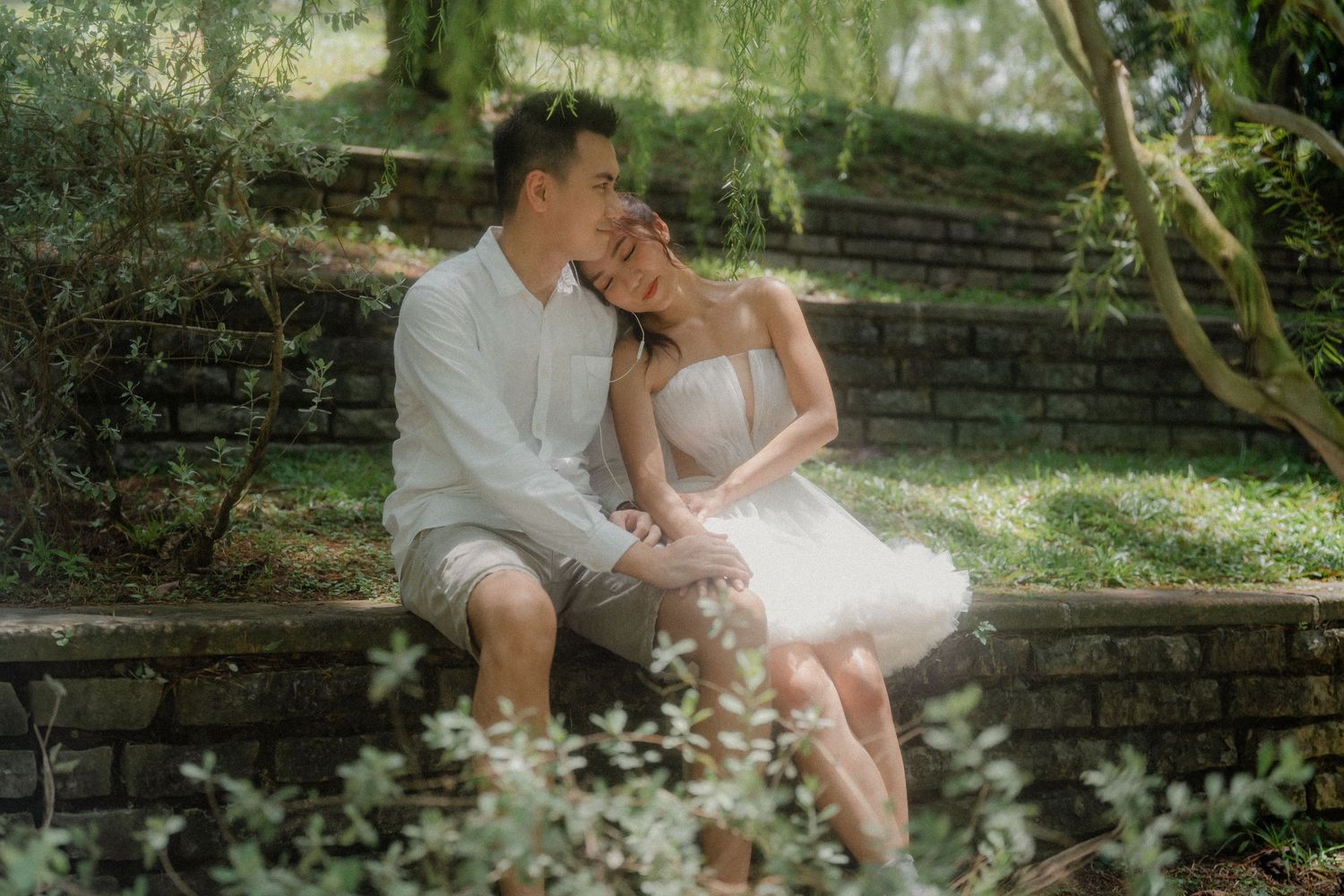 A couple embraces intimately on a rustic stone bench surrounded by lush greenery in a garden setting.