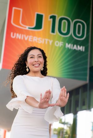 Joyful woman in white dress poses with outstretched hands in front of University of Miami 100 sign.