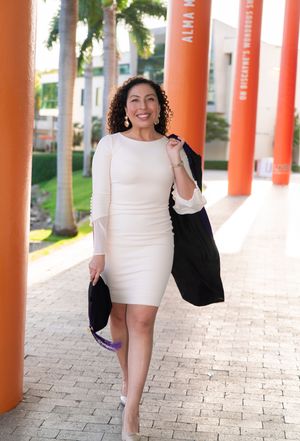 Elegant woman in white dress carries black graduation gown while walking between orange campus columns.