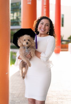Smiling woman in white dress holds a cute golden doodle puppy wearing a graduation cap.