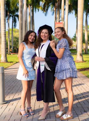 Graduate in black gown and cap smiles with two young women on a sunny campus path.