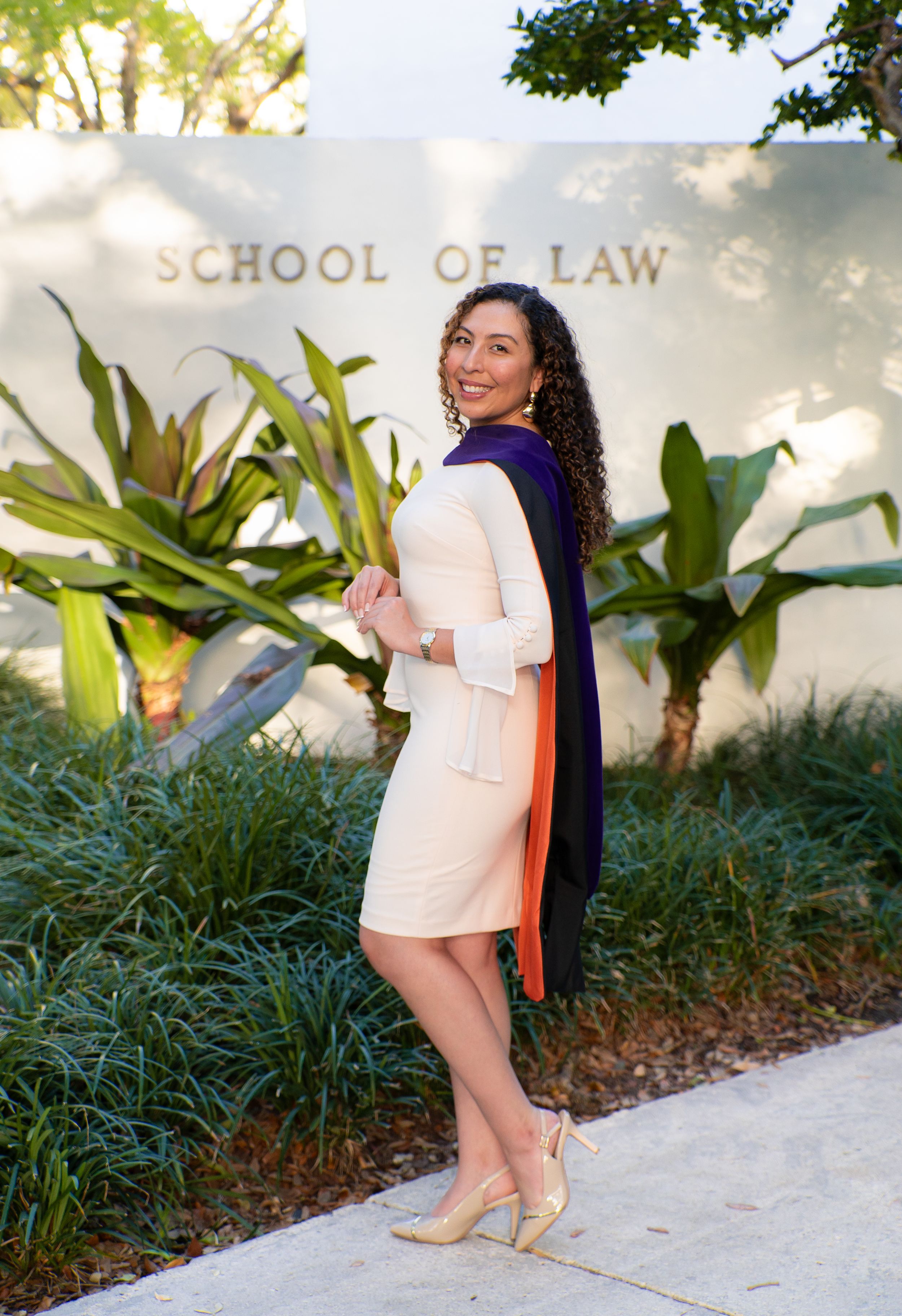 Law school graduate smiles in cream dress with purple and orange academic hood outside School of Law building.