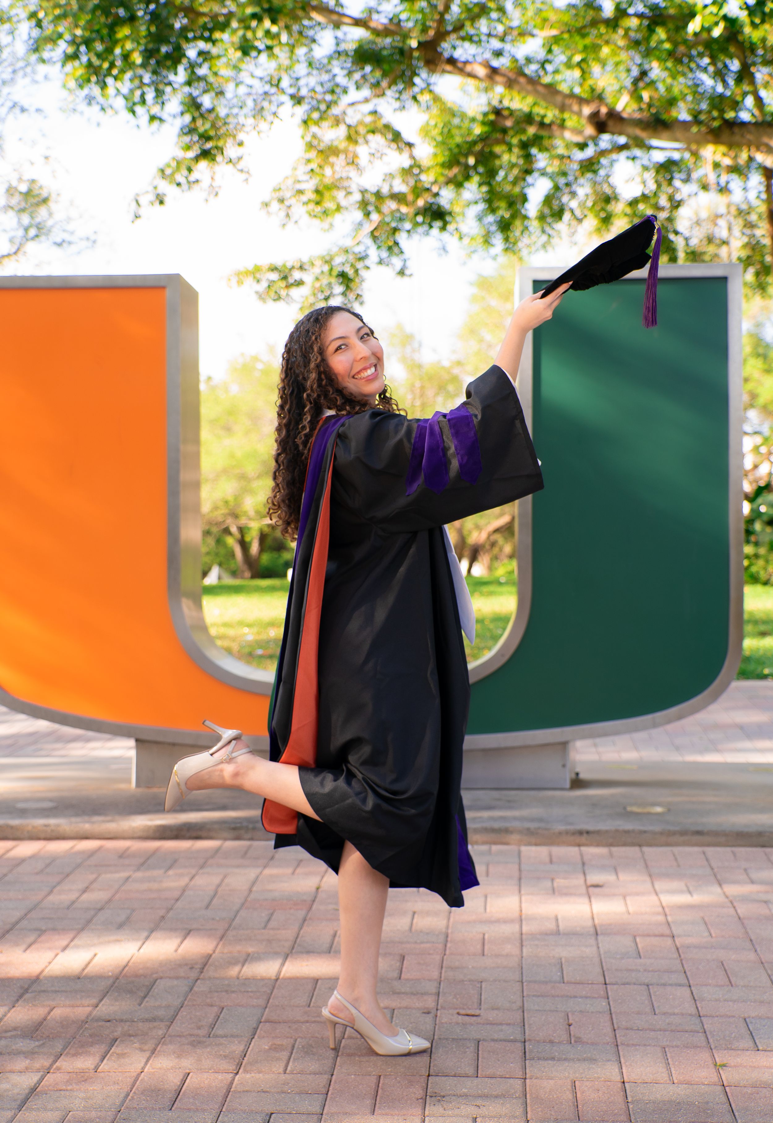 Smiling graduate in black gown tosses cap beside colorful University of Miami U sculpture.