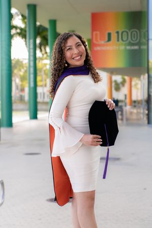 Smiling woman in white dress holds graduation cap outside University of Miami 100th anniversary banner.