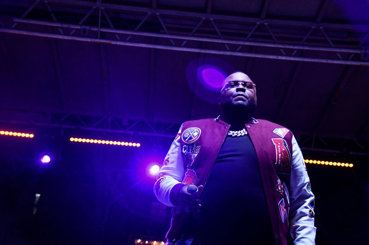 A performer in a varsity jacket energetically sings on stage under dramatic purple lighting during a concert performance.