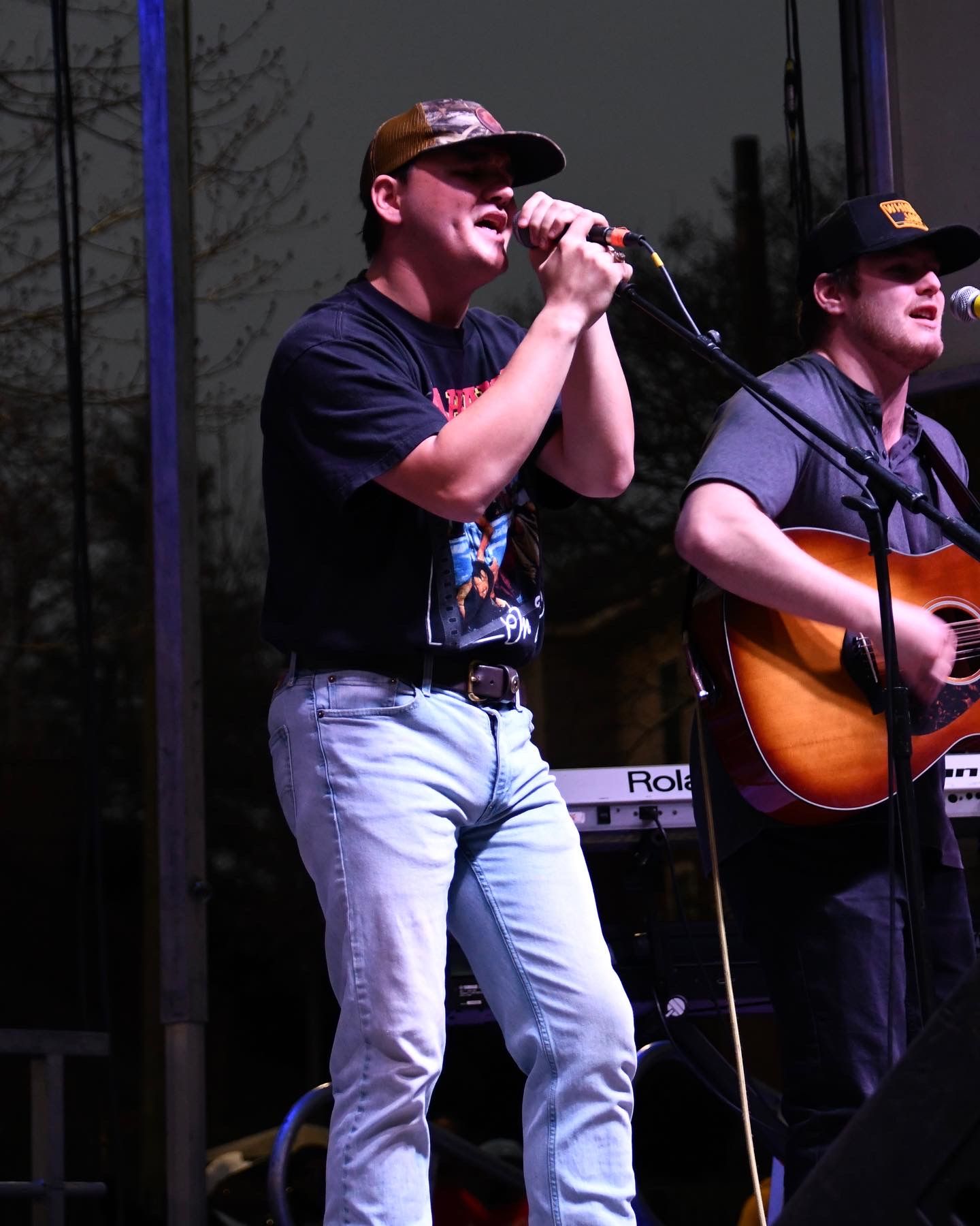 Musicians perform on stage with acoustic guitar and microphone during an outdoor concert at dusk.