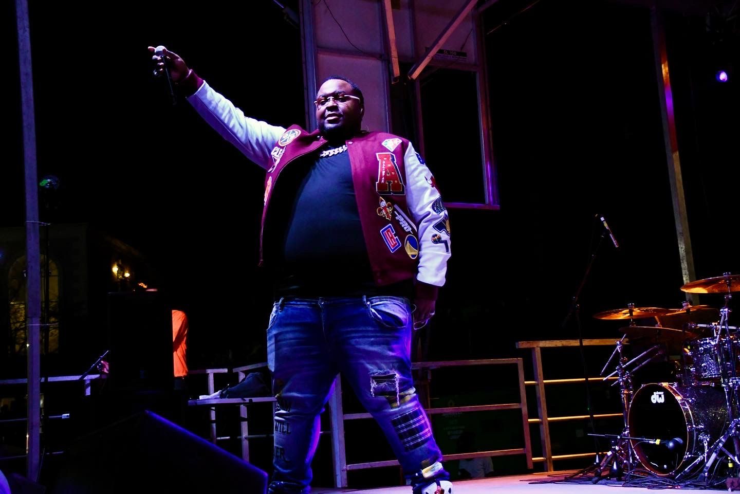 Performer in red vest and jeans sings passionately on dimly lit stage with drum kit visible in background.