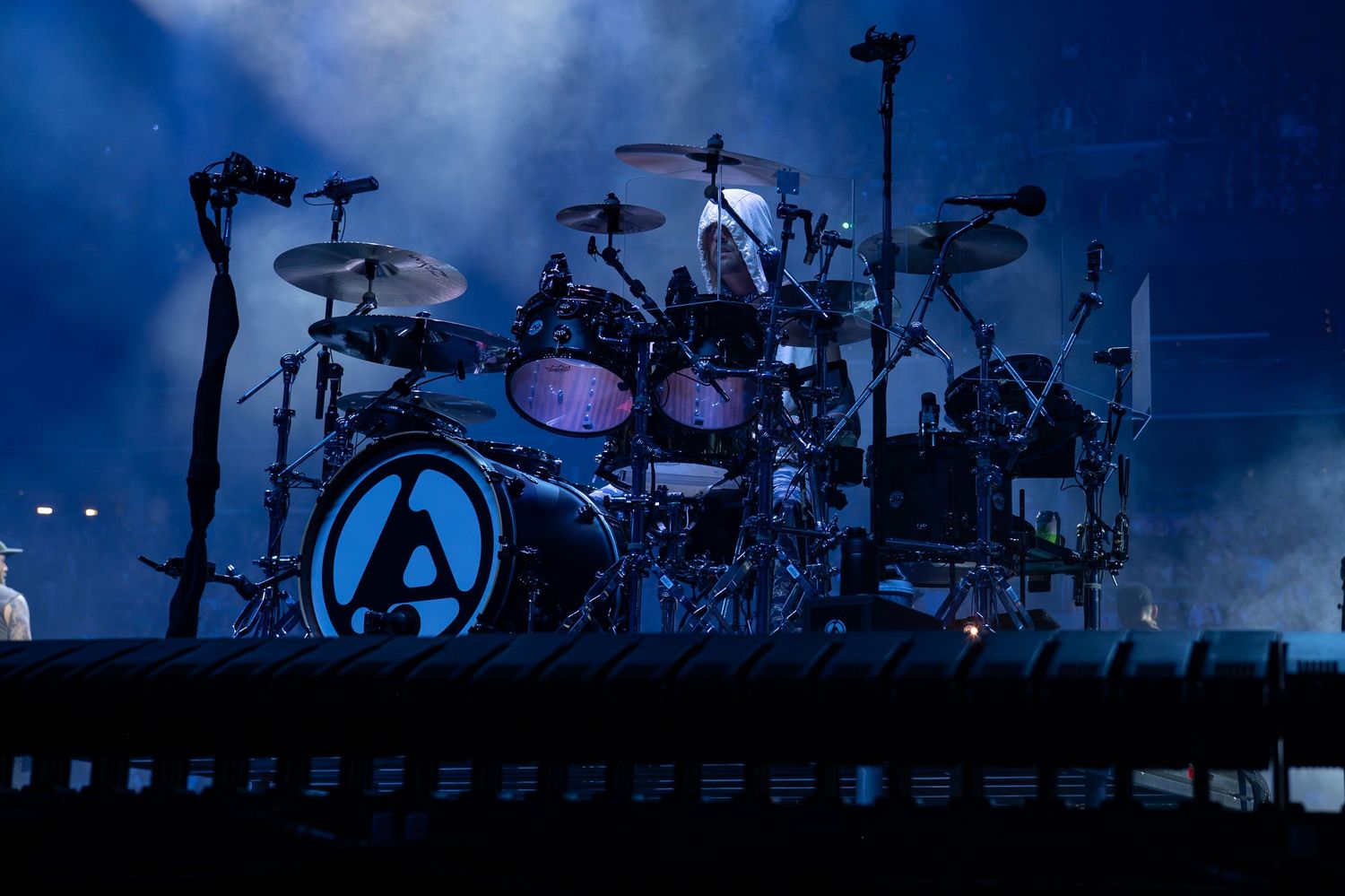 Silhouettes of drum kits and musical equipment on stage against a moody blue-lit concert backdrop.