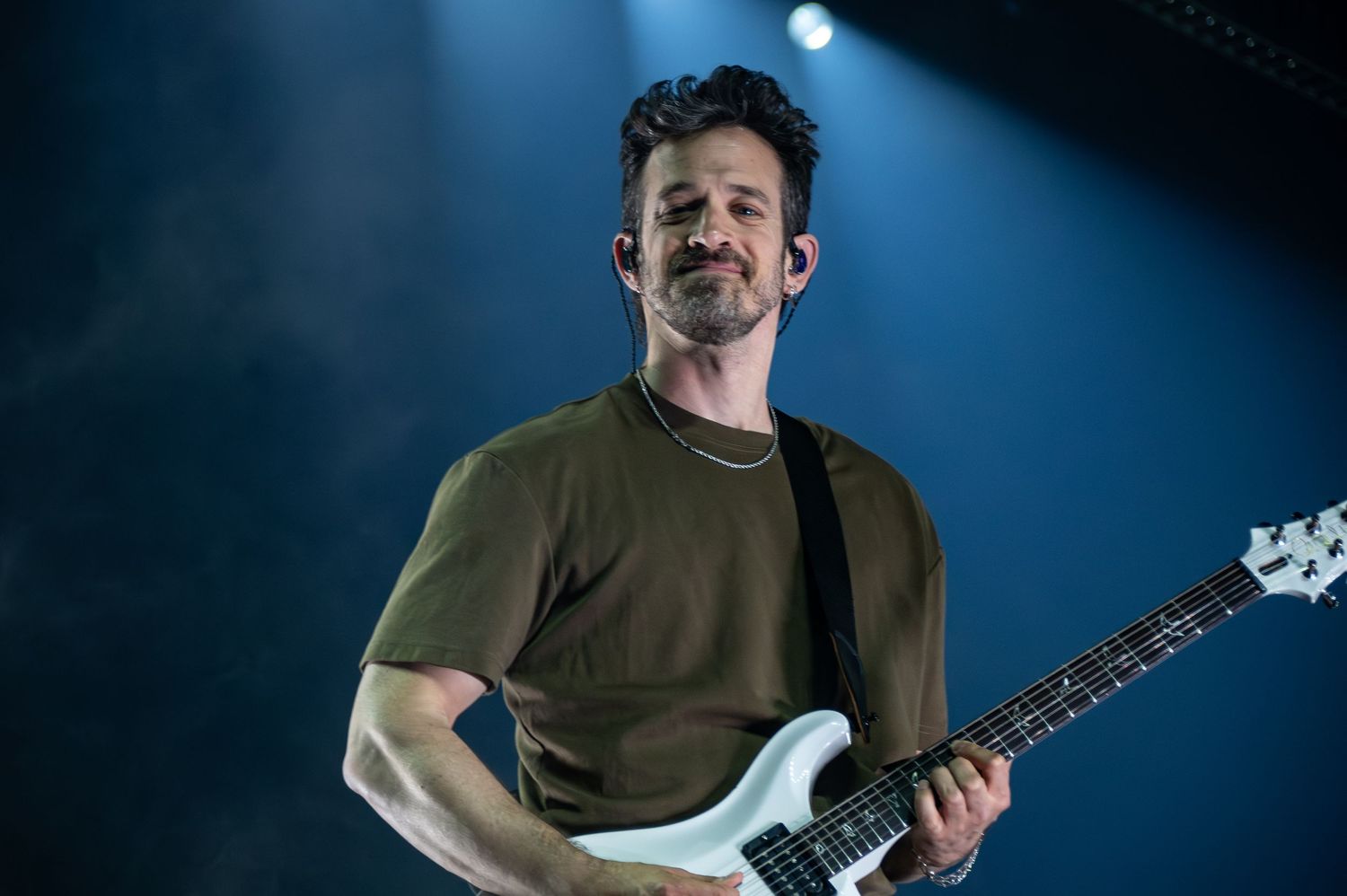 A musician plays an electric guitar on a dark stage with blue lighting effects in the background.