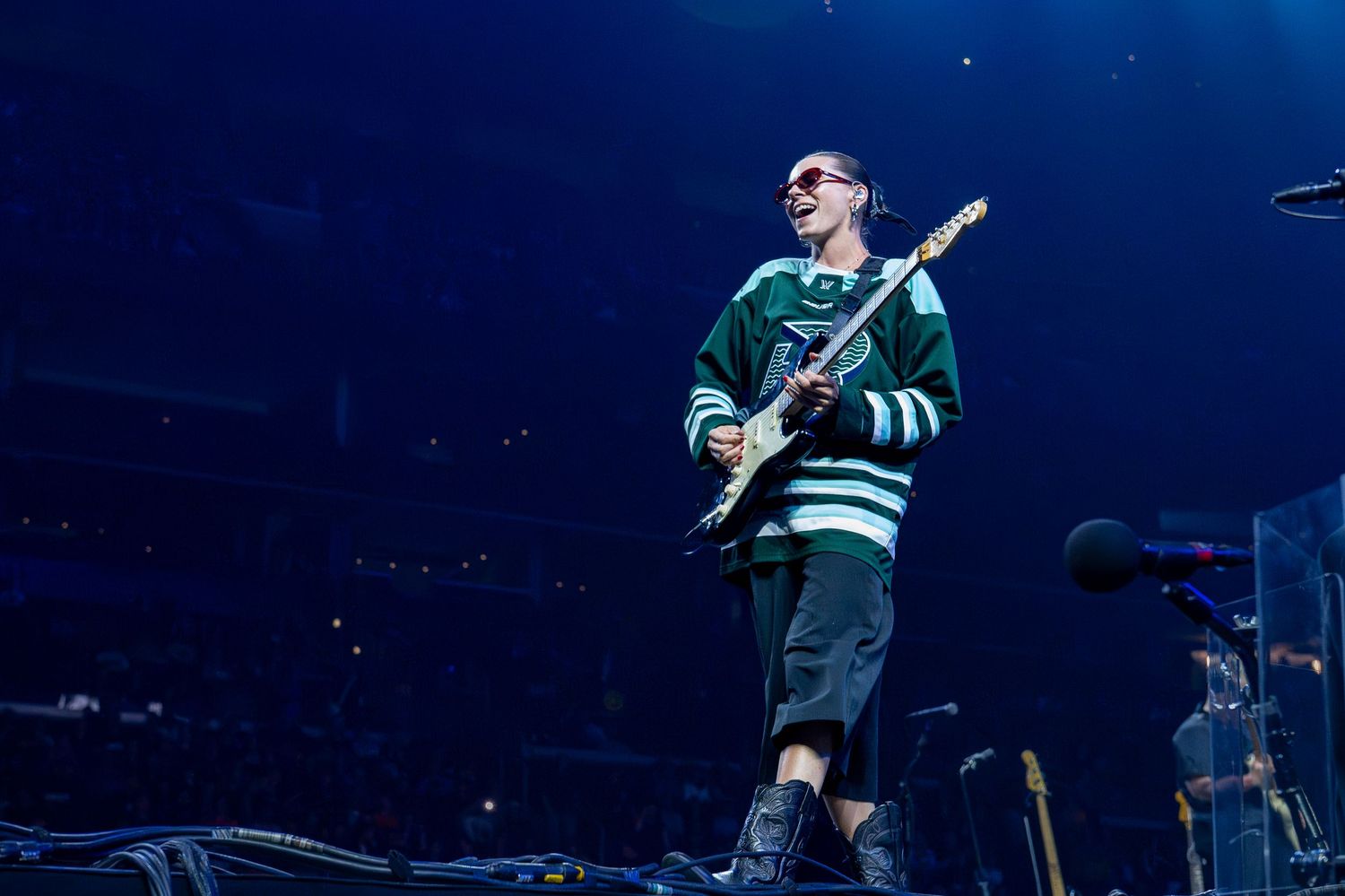 A musician performs on stage wearing a green and white striped jersey while playing an electric guitar under blue lighting.
