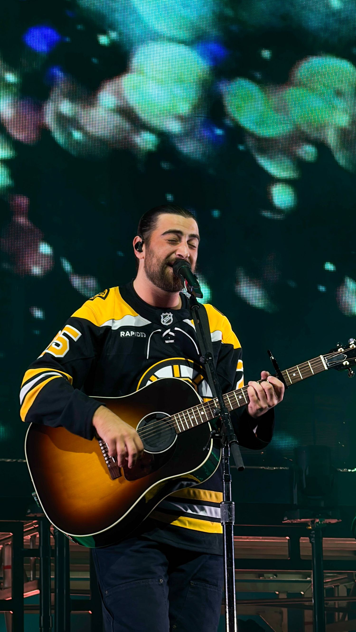 A musician wearing a Boston Bruins hockey jersey performs on an acoustic guitar against a colorful illuminated backdrop.