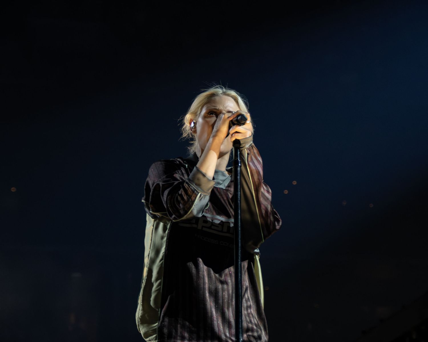 A person in dark clothing stands against a black background with dramatic lighting and shadows.