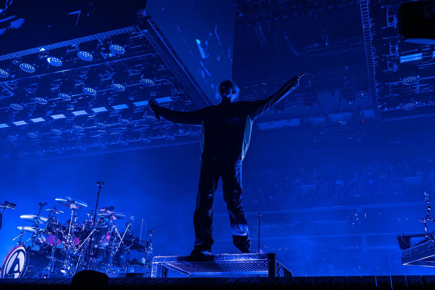 A silhouetted performer stands with arms outstretched on stage bathed in dramatic blue lighting during a concert performance.