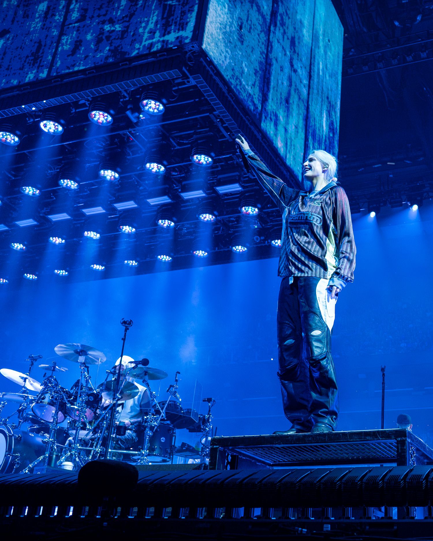 A performer stands on stage bathed in blue lighting with dramatic overhead stage lights and drum kit visible in the background.
