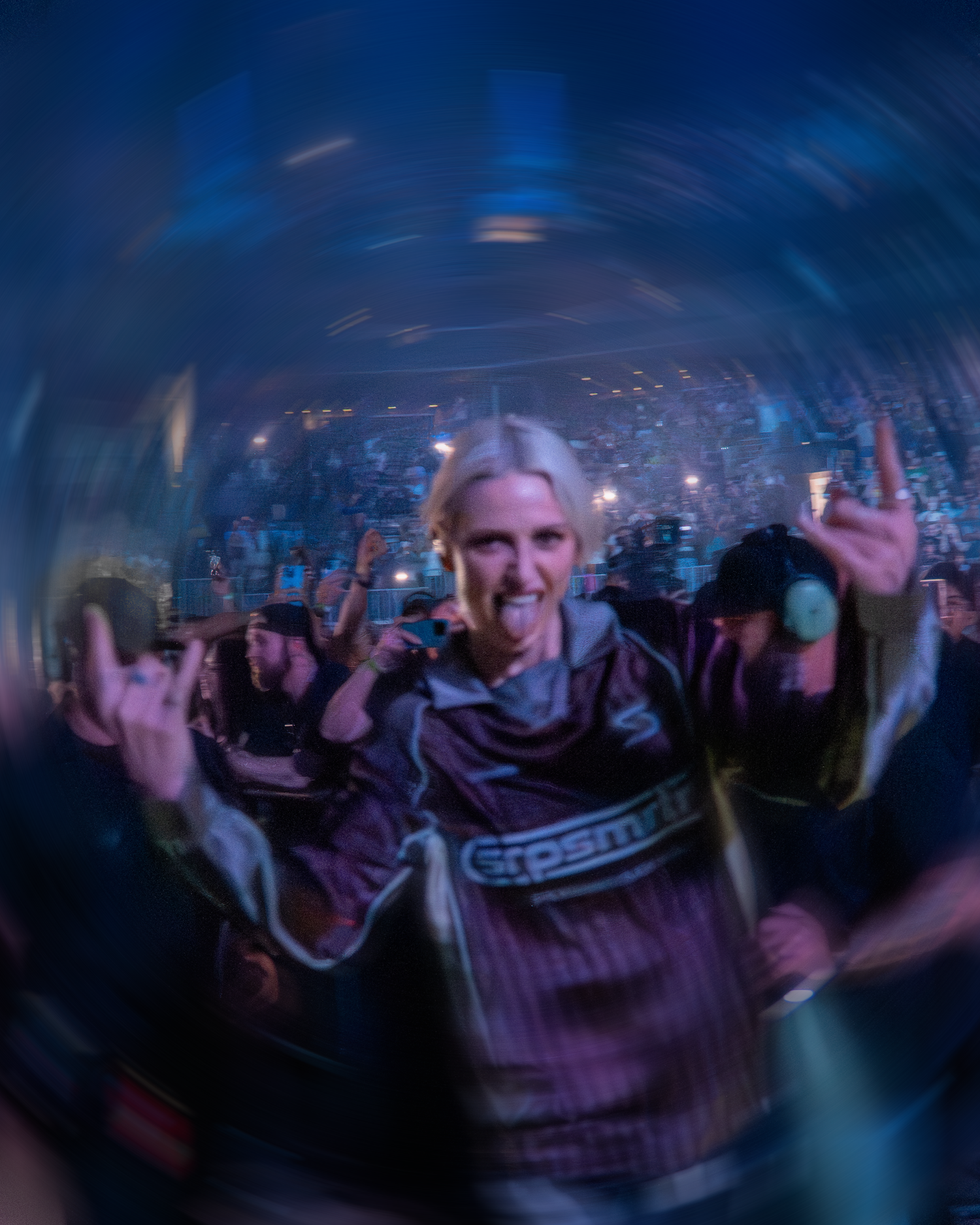 Crowd of concert-goers with raised hands dancing in a dark nightclub with blue lighting and atmospheric effects.
