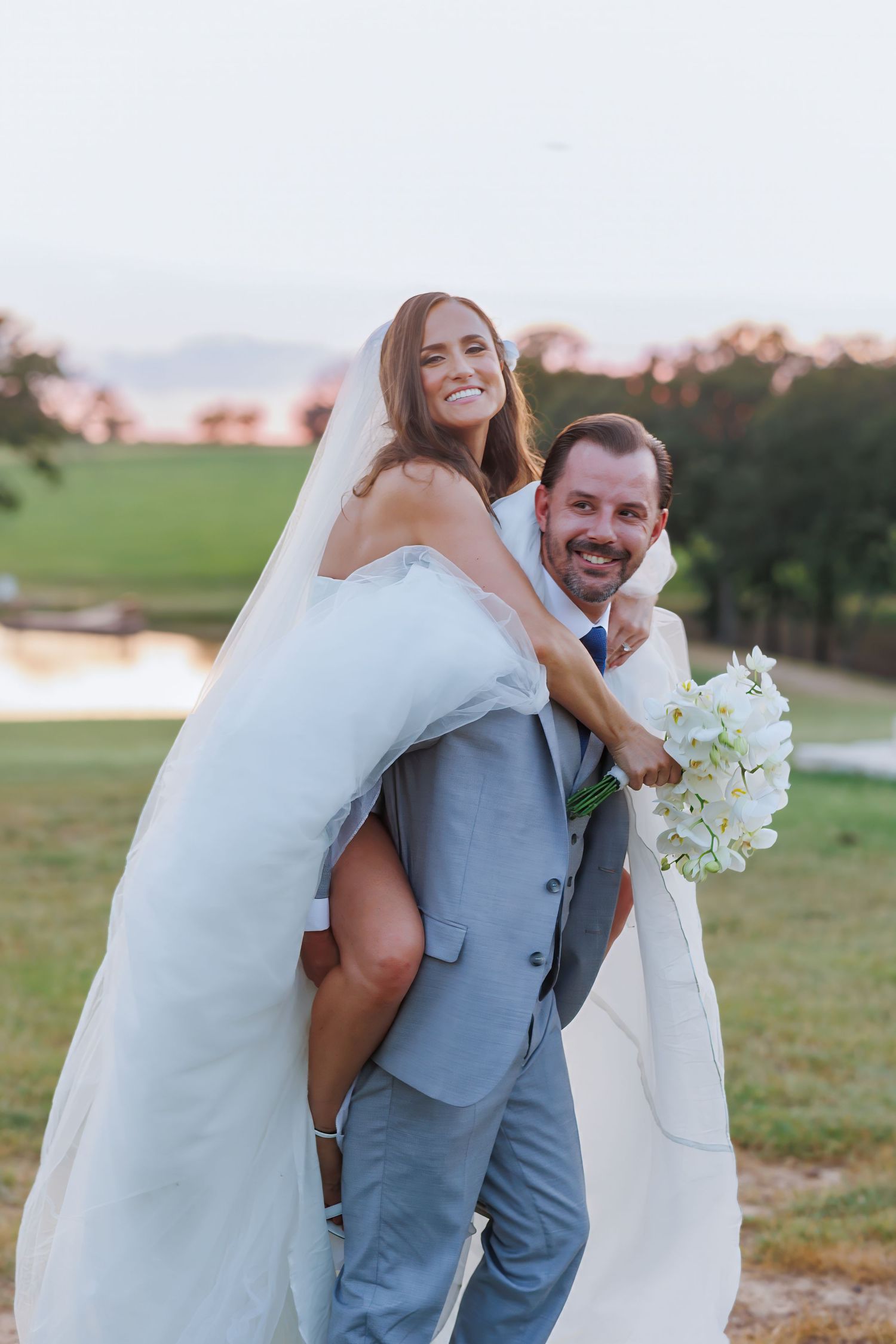 Joyful couple in wedding attire sharing a playful piggyback moment at sunset on the lawn at Bella Cavalli