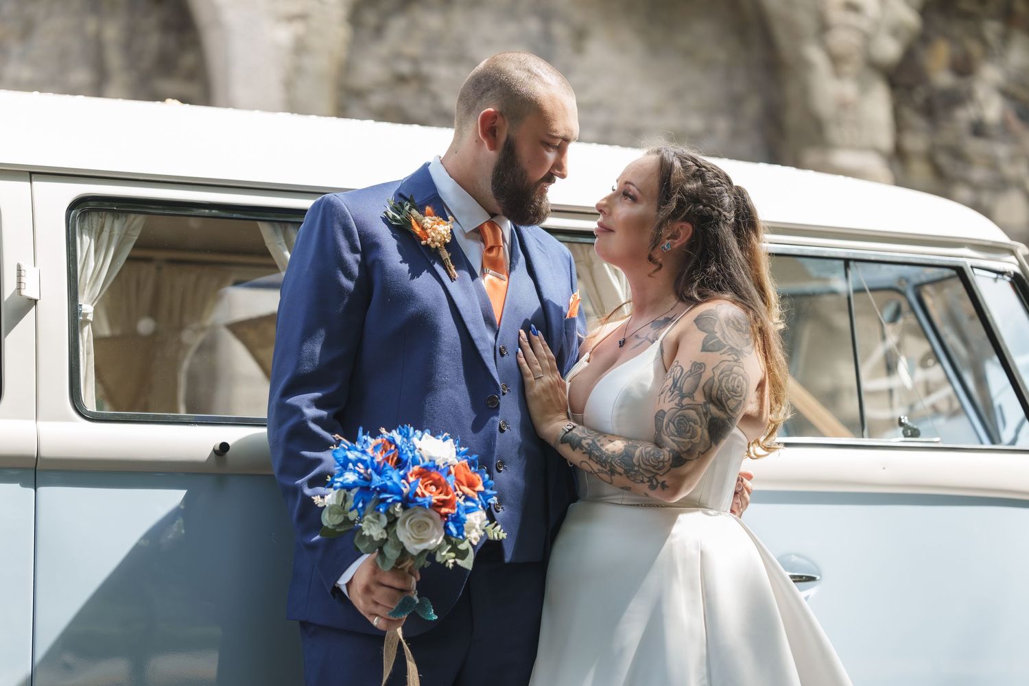 A newlywed couple poses beside a classic VW bus with blue floral bouquet.