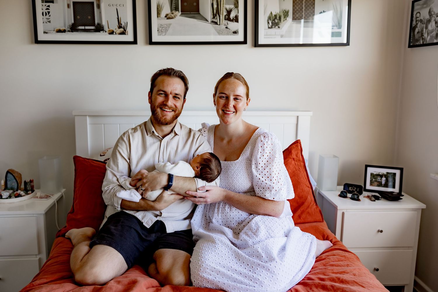Two adults sitting on a red bed with their newborn twins in a bright bedroom with framed artwork on the wall.
