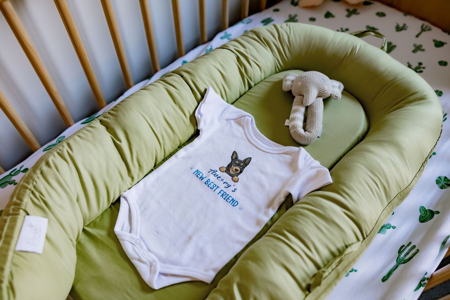 A white baby onesie with a graphic design lies on a green cushioned baby nest with a small plush toy.