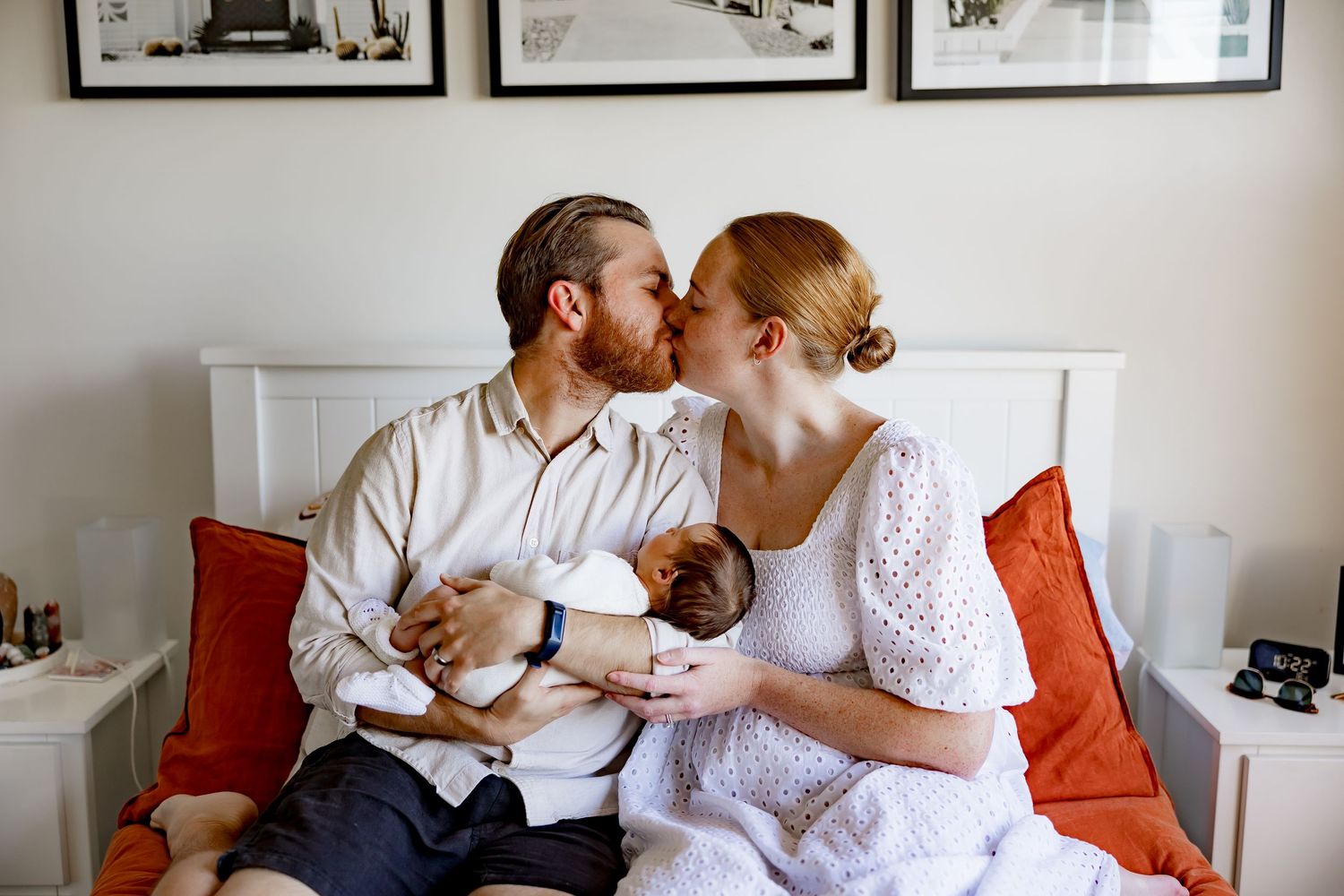 A couple shares an intimate moment while lounging on a white couch with orange pillows against a wall with framed artwork.