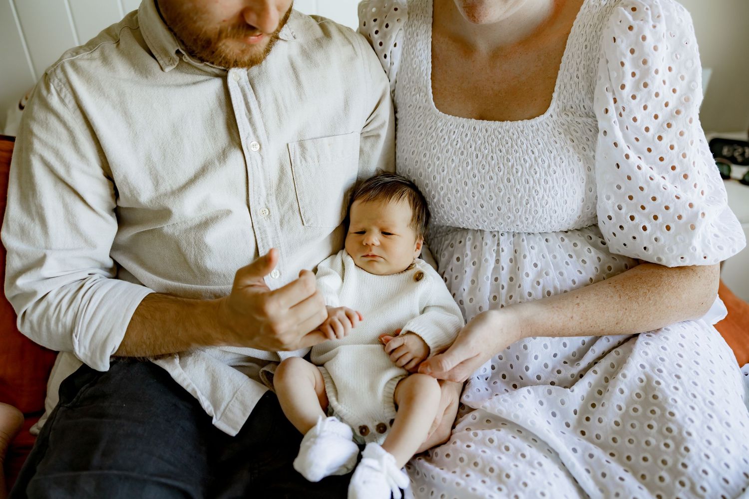Sleeping newborn infant in white onesie held by parents wearing white outfits while sitting together.