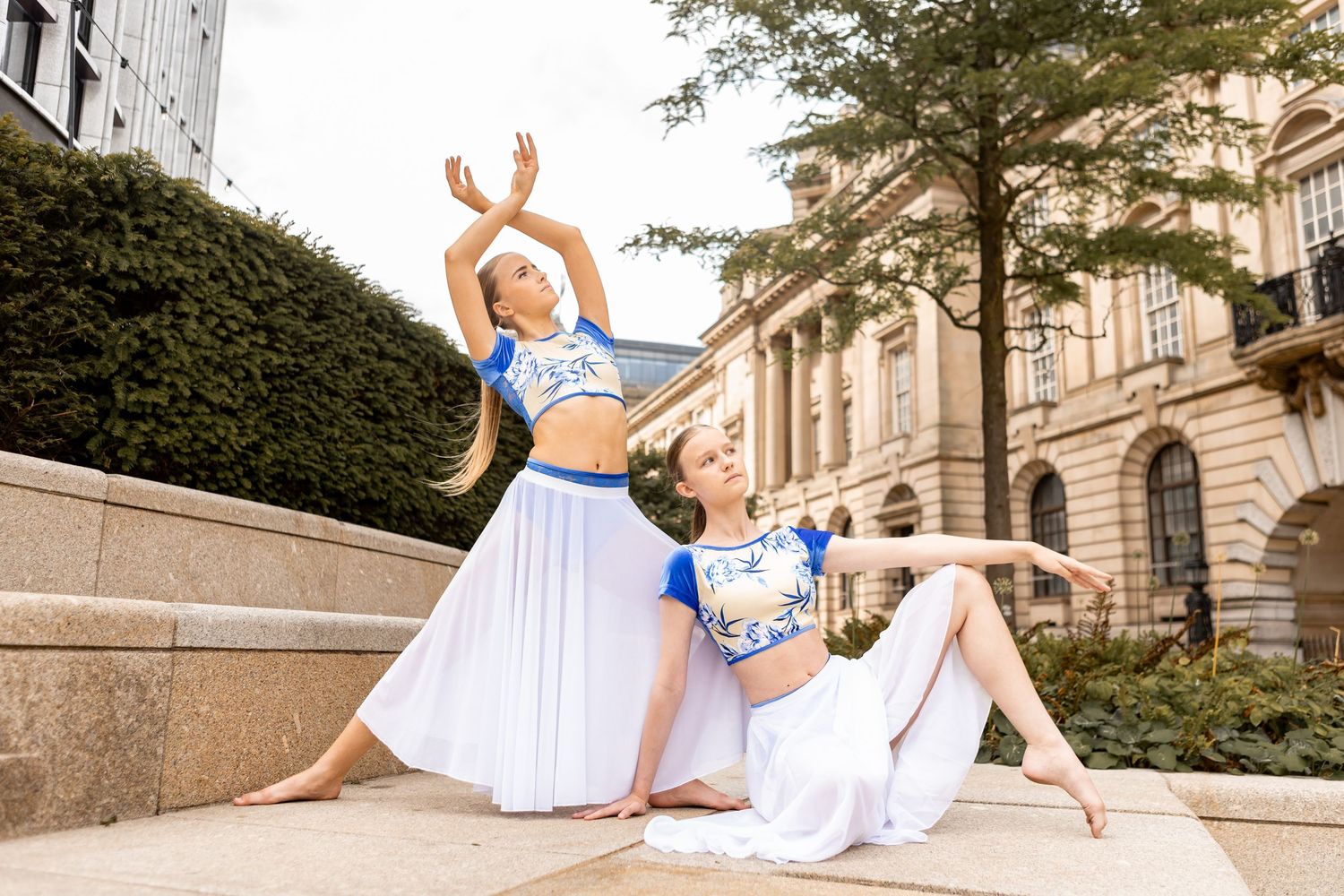Two ballet dancers in flowing white dresses perform graceful poses on stone steps outside an elegant building.