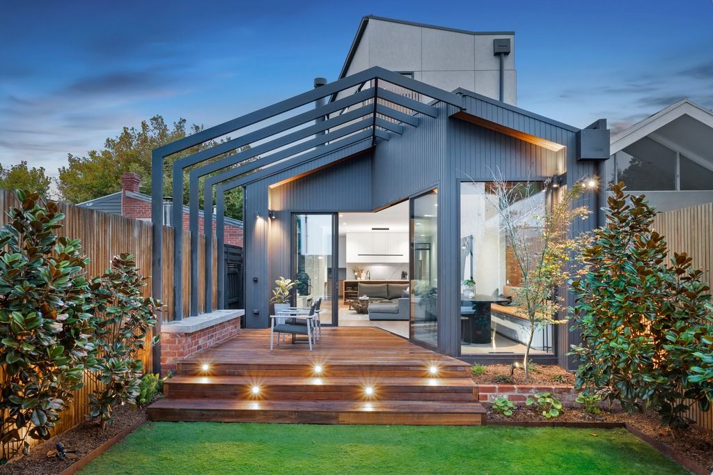 Modern backyard deck with pergola features integrated lighting and landscaping on a contemporary blue-gray house exterior.