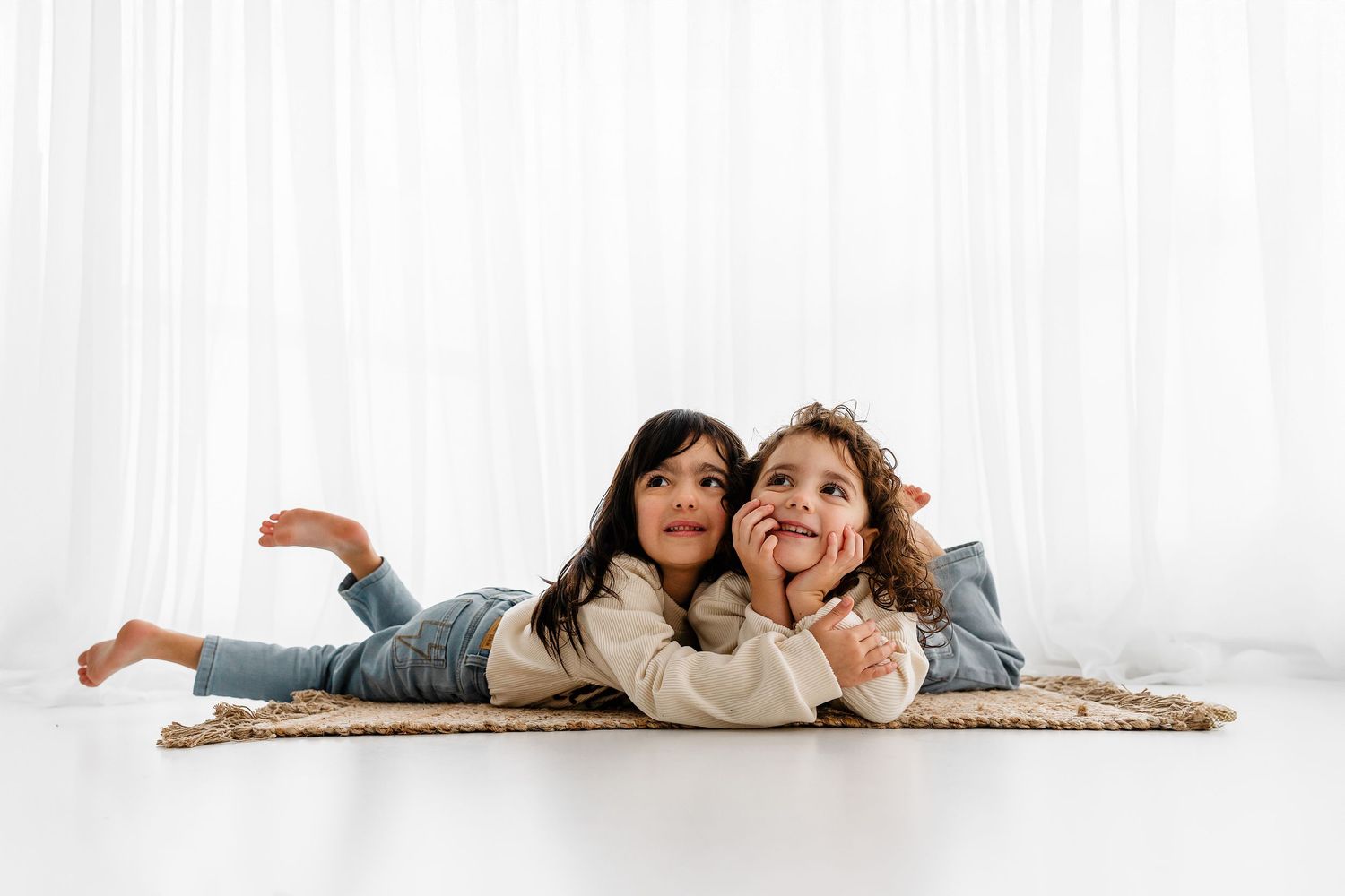 Two children in denim lying down together on a white carpet near a white curtain background.