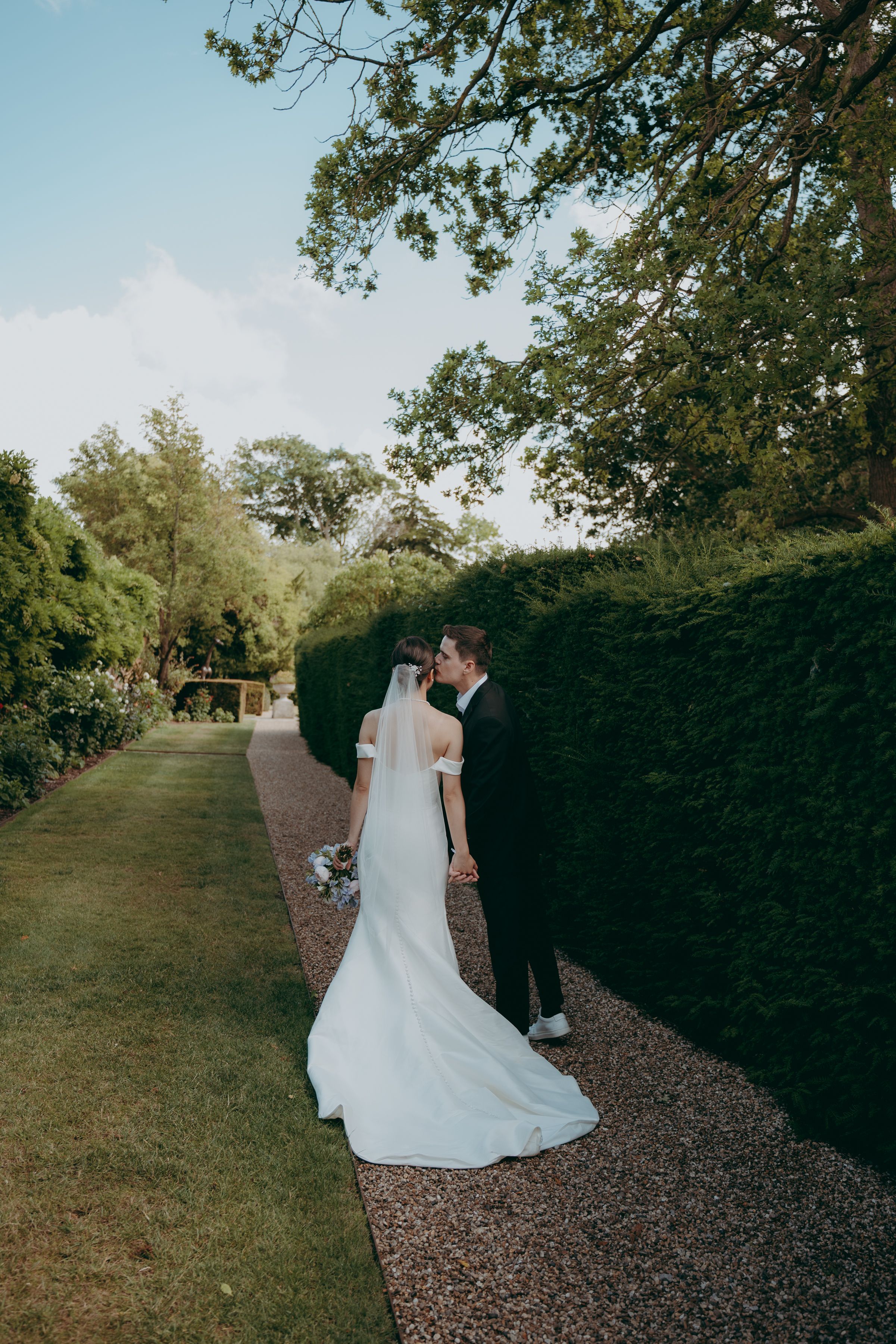 Newlyweds walking together on a gravel path lined with green trees and hedges on a sunny day.