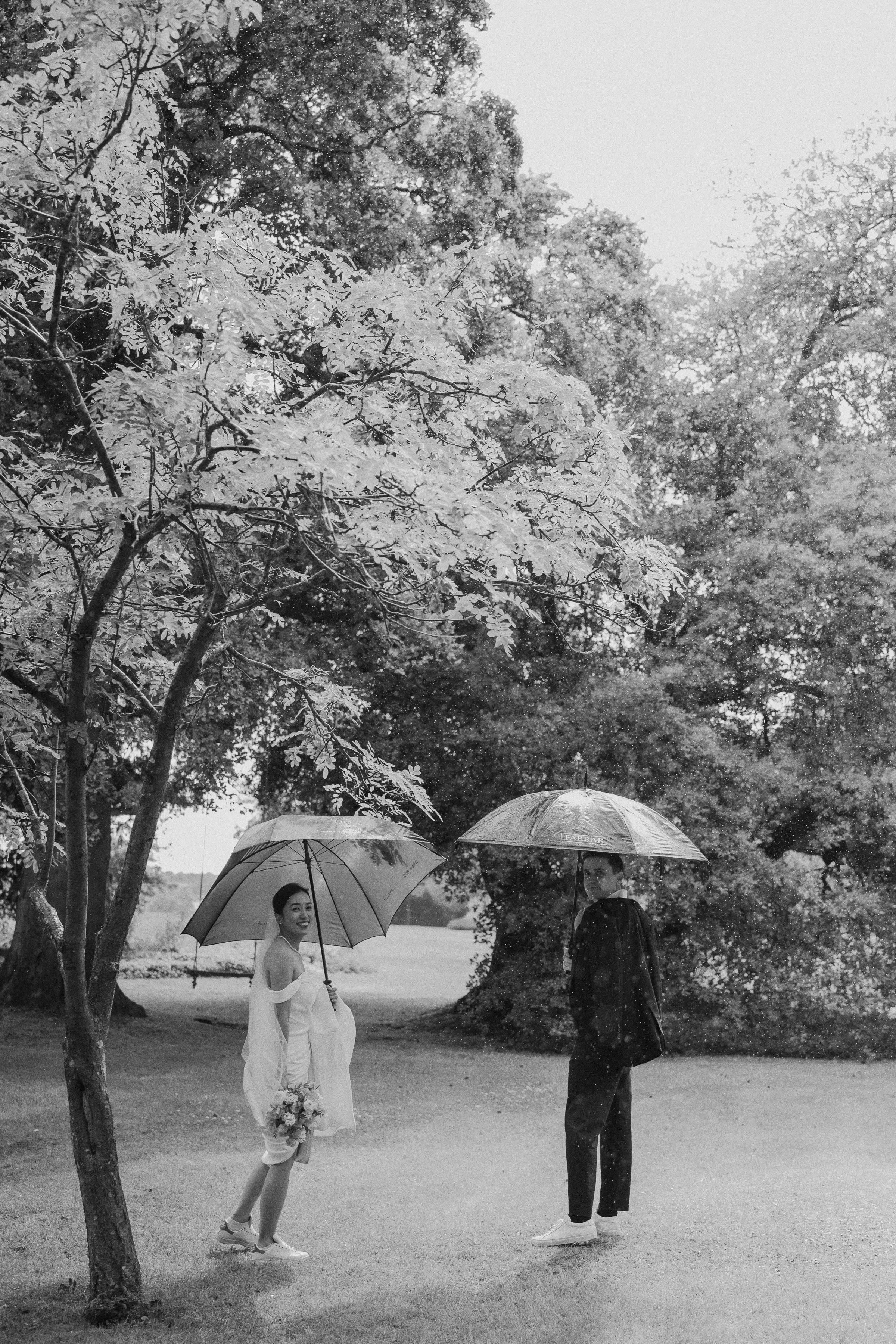 Two people holding umbrellas stand beneath blossoming trees in a dreamy black and white garden scene.
