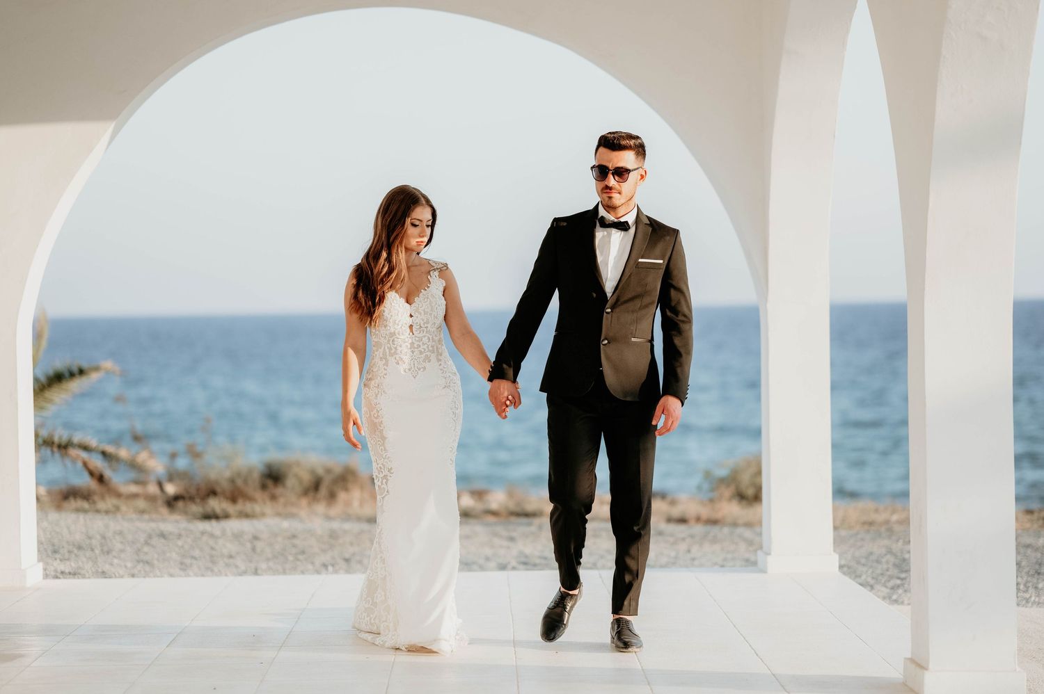 Two people in elegant wedding attire walk together along a scenic coastal beach at sunset.