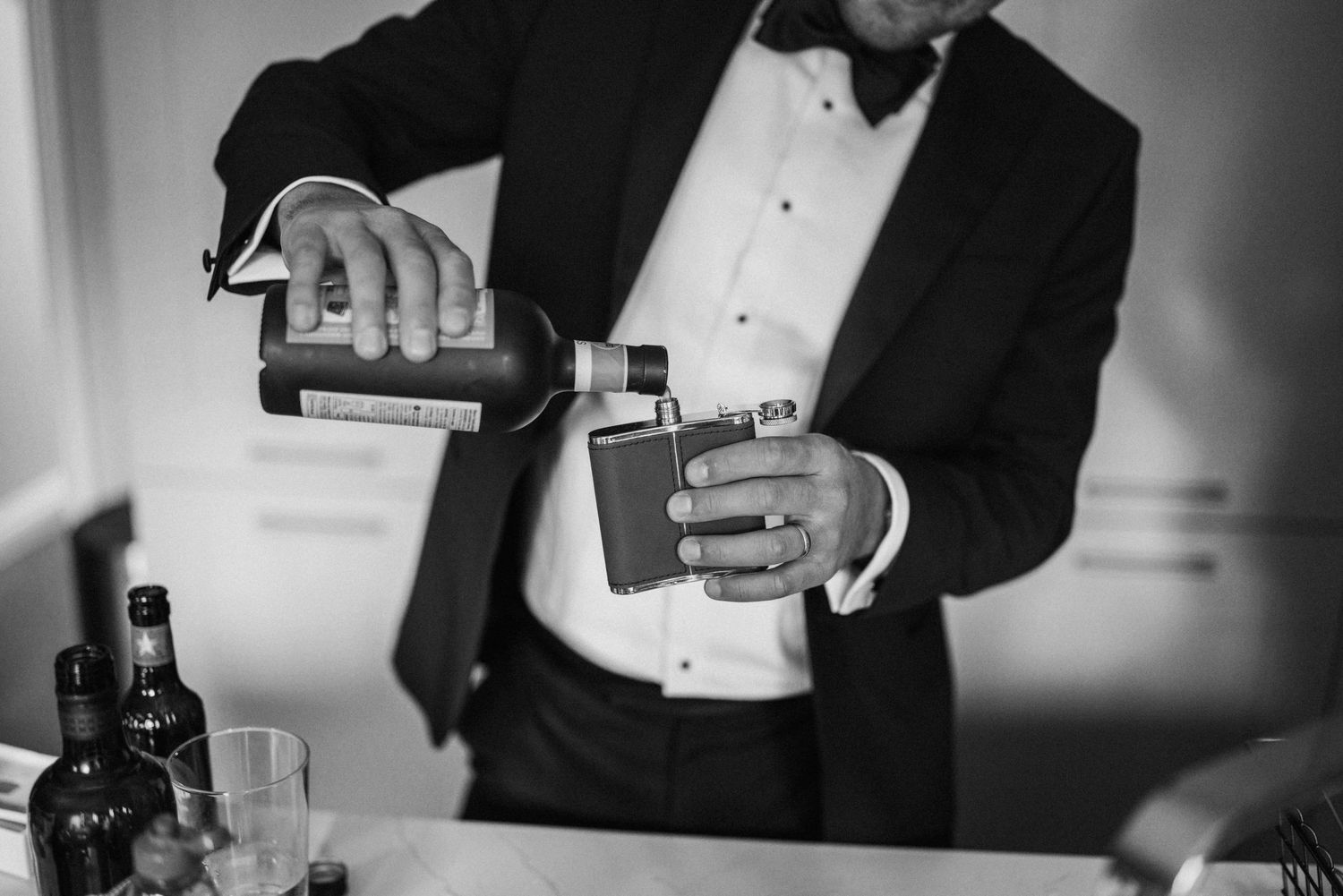Black and white photo of someone in formal wear pouring drinks at a bar, with bottles in the background.