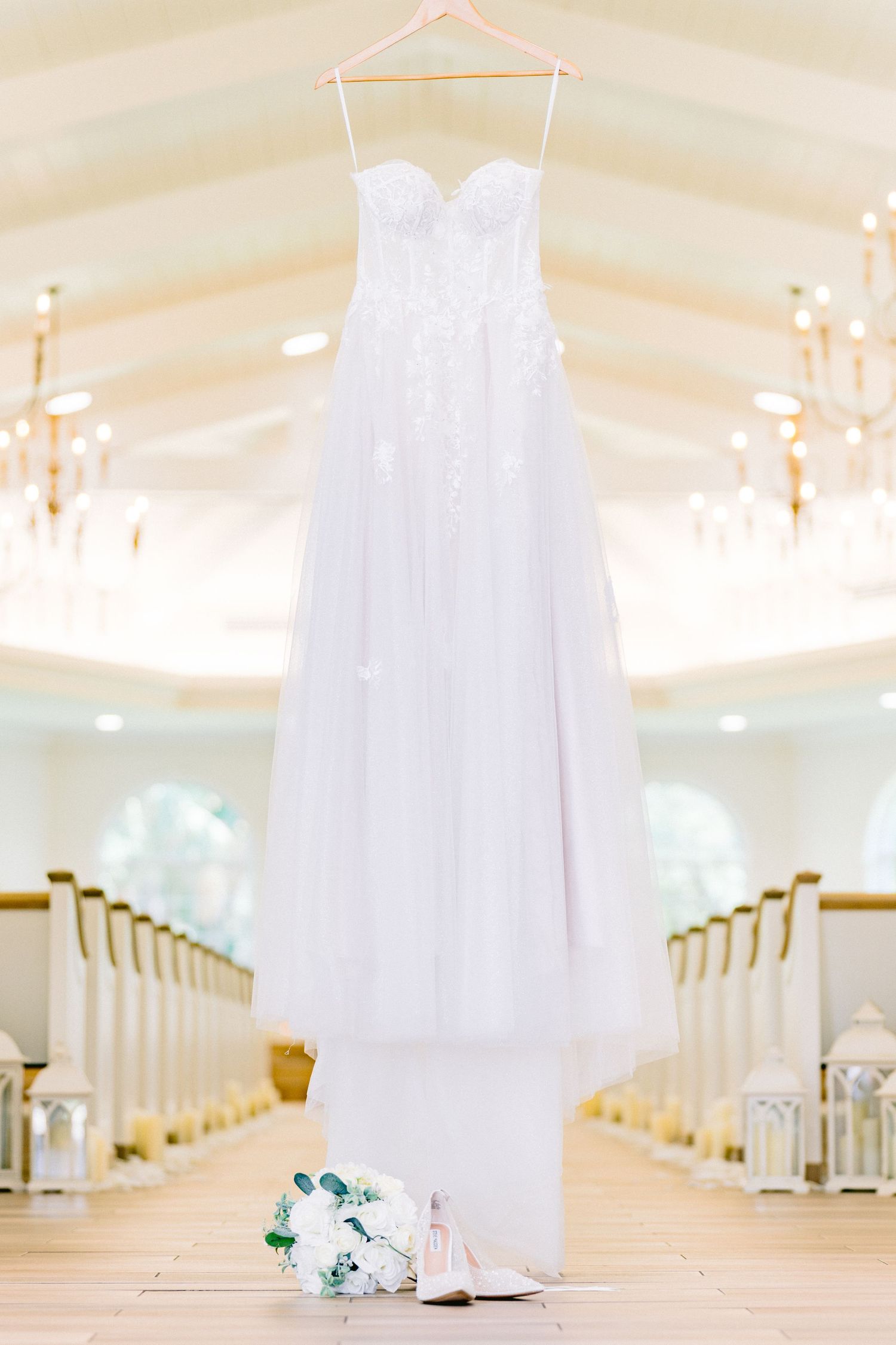 Elegant white wedding dress displayed in a light-filled venue, Harborside Chapel, with rows of white chairs and chandeliers overhead.
