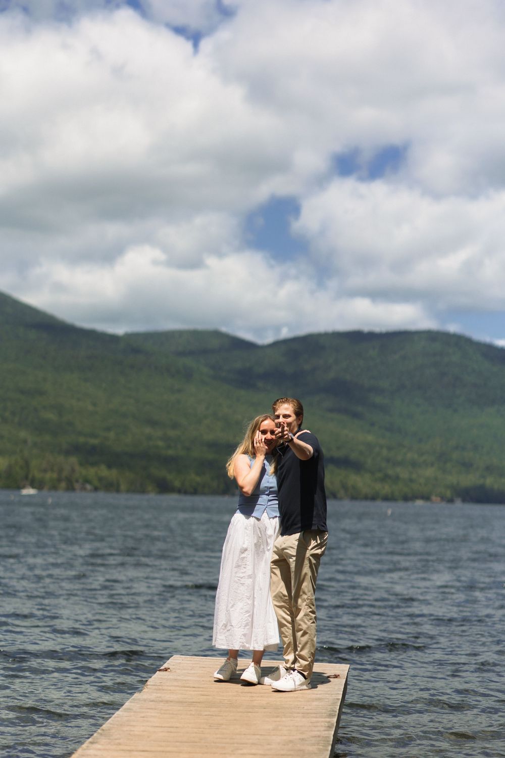Two people stand on a wooden dock overlooking a scenic mountain lake with cloudy skies above.