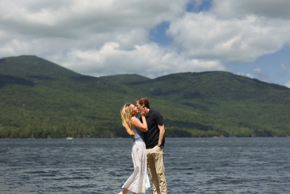 A couple shares a romantic kiss on a boat deck with mountain lake scenery and cloudy skies in the background.