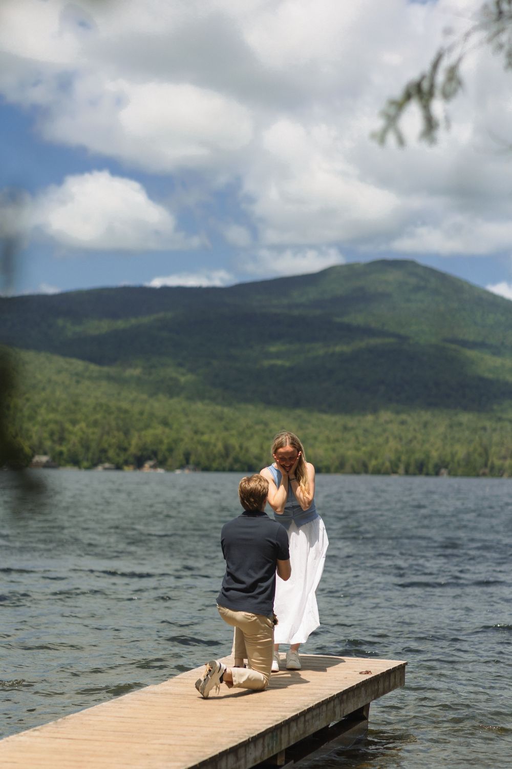 A romantic proposal moment unfolds on a wooden dock overlooking a pristine mountain lake on a beautiful summer day.