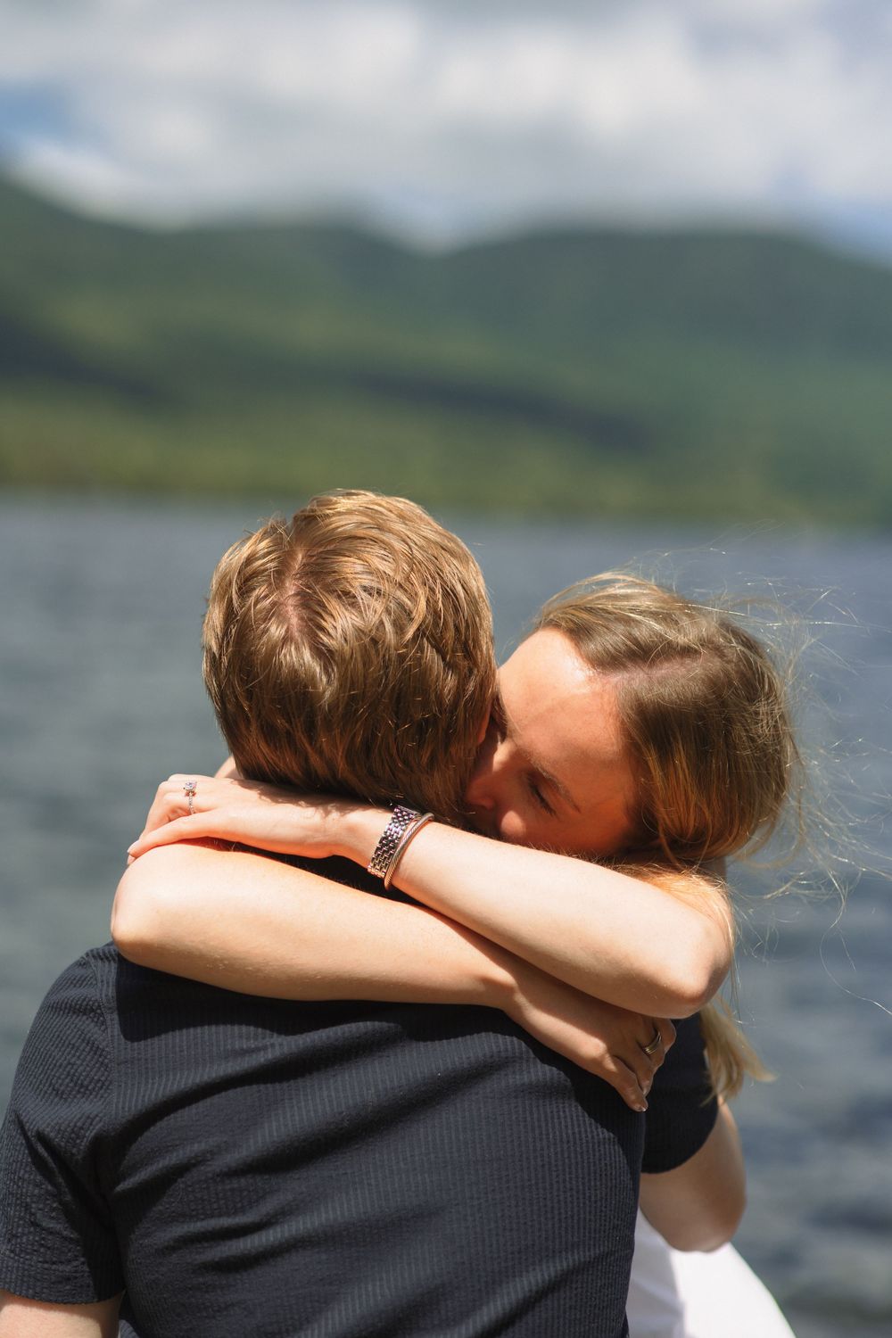 Two people embrace in a tender moment by a scenic lake with mountains in the background.