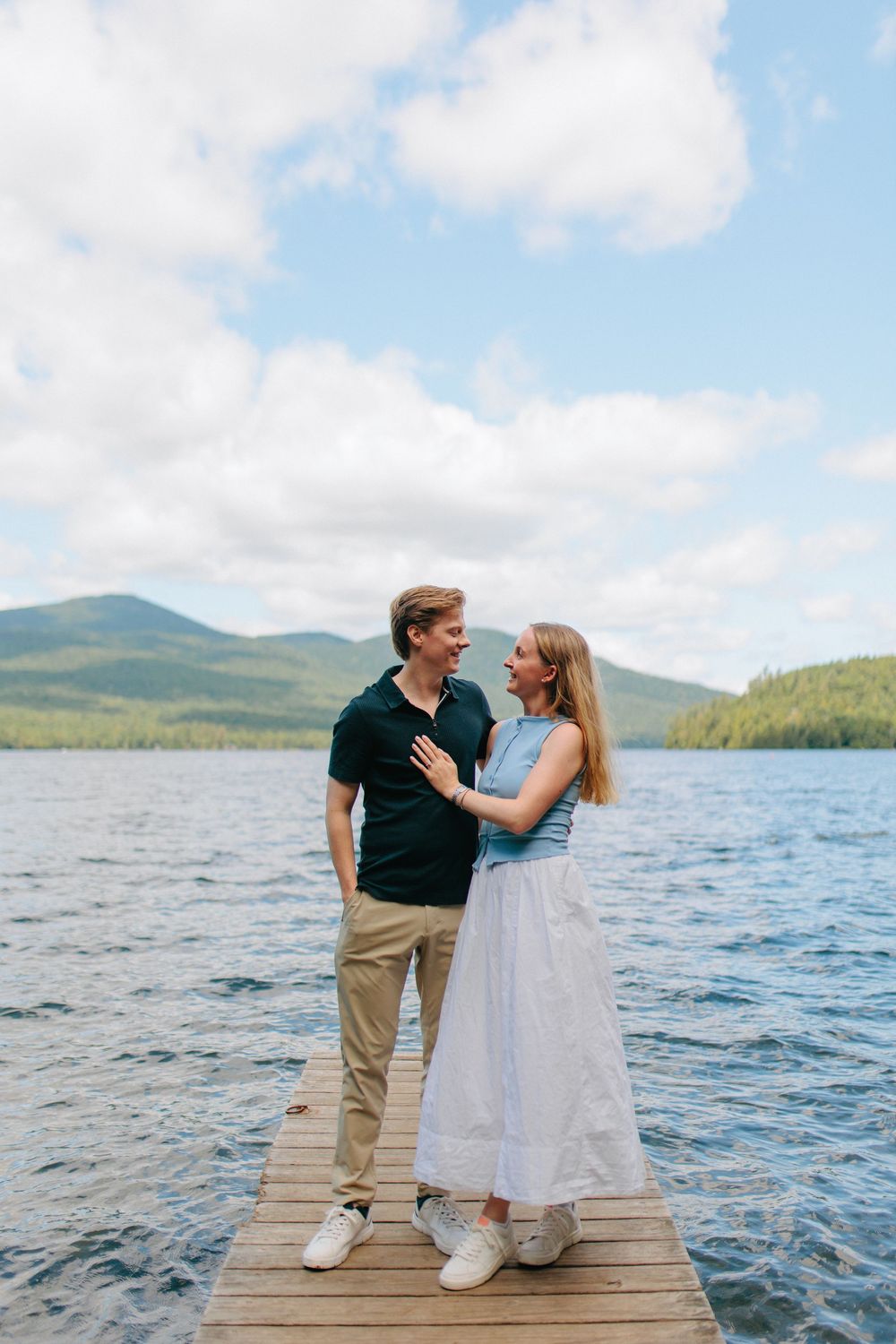 A couple shares a romantic moment on a wooden dock overlooking a lake surrounded by mountains on a sunny day.