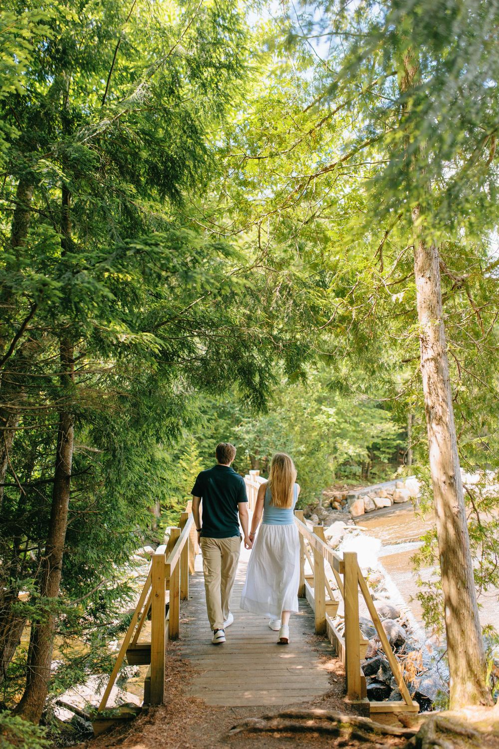 A couple walks hand in hand across a wooden footbridge in a lush green forest setting.