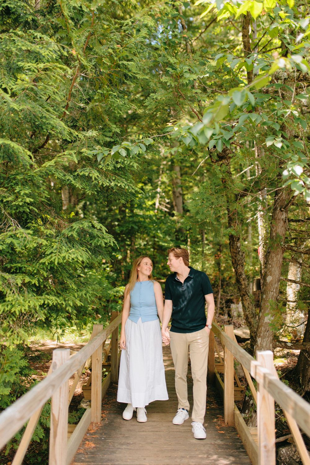 A couple walks together on a wooden footbridge surrounded by lush green trees and foliage in a peaceful forest setting.