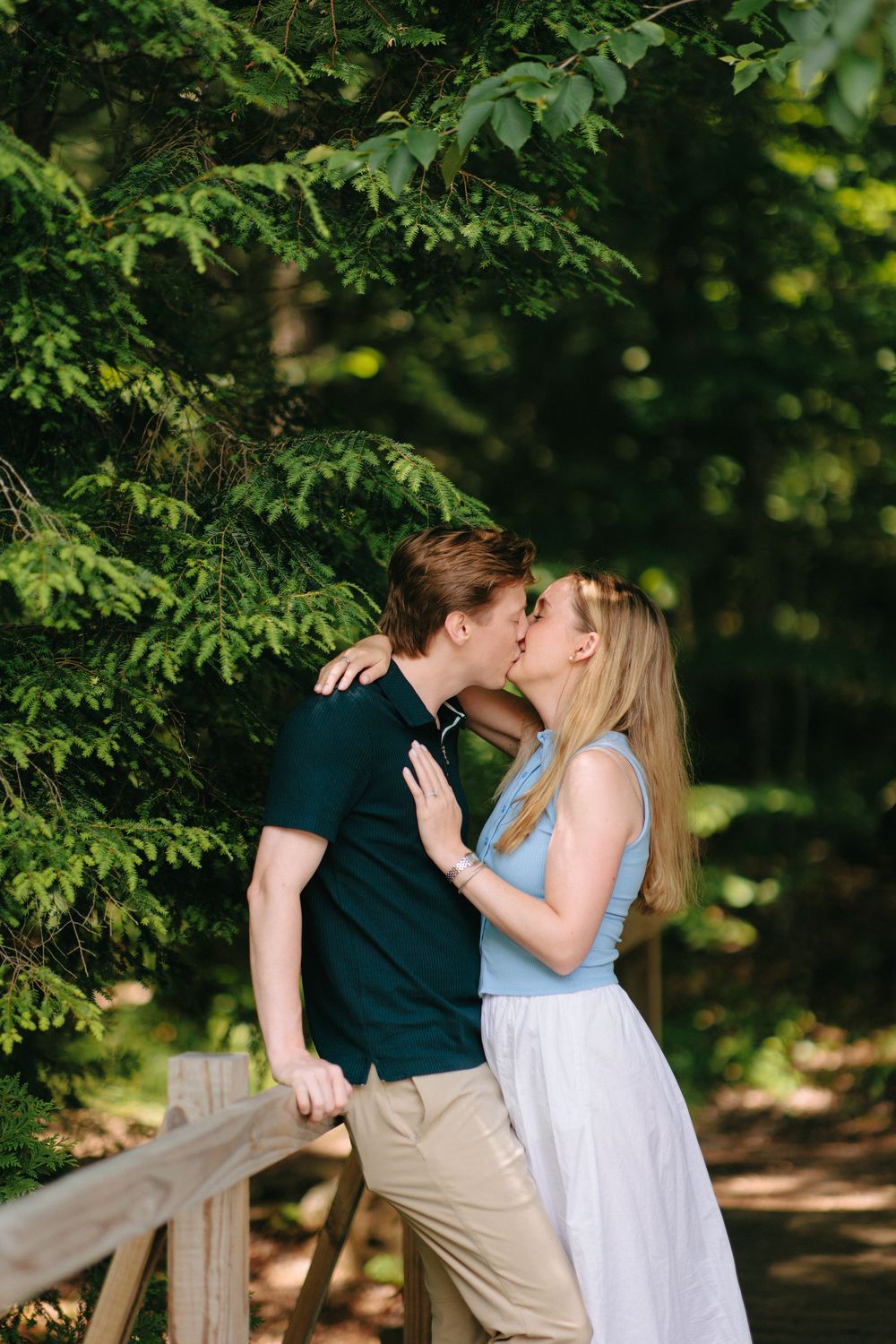 A couple in casual summer attire shares a romantic moment on a wooden deck surrounded by lush green trees.