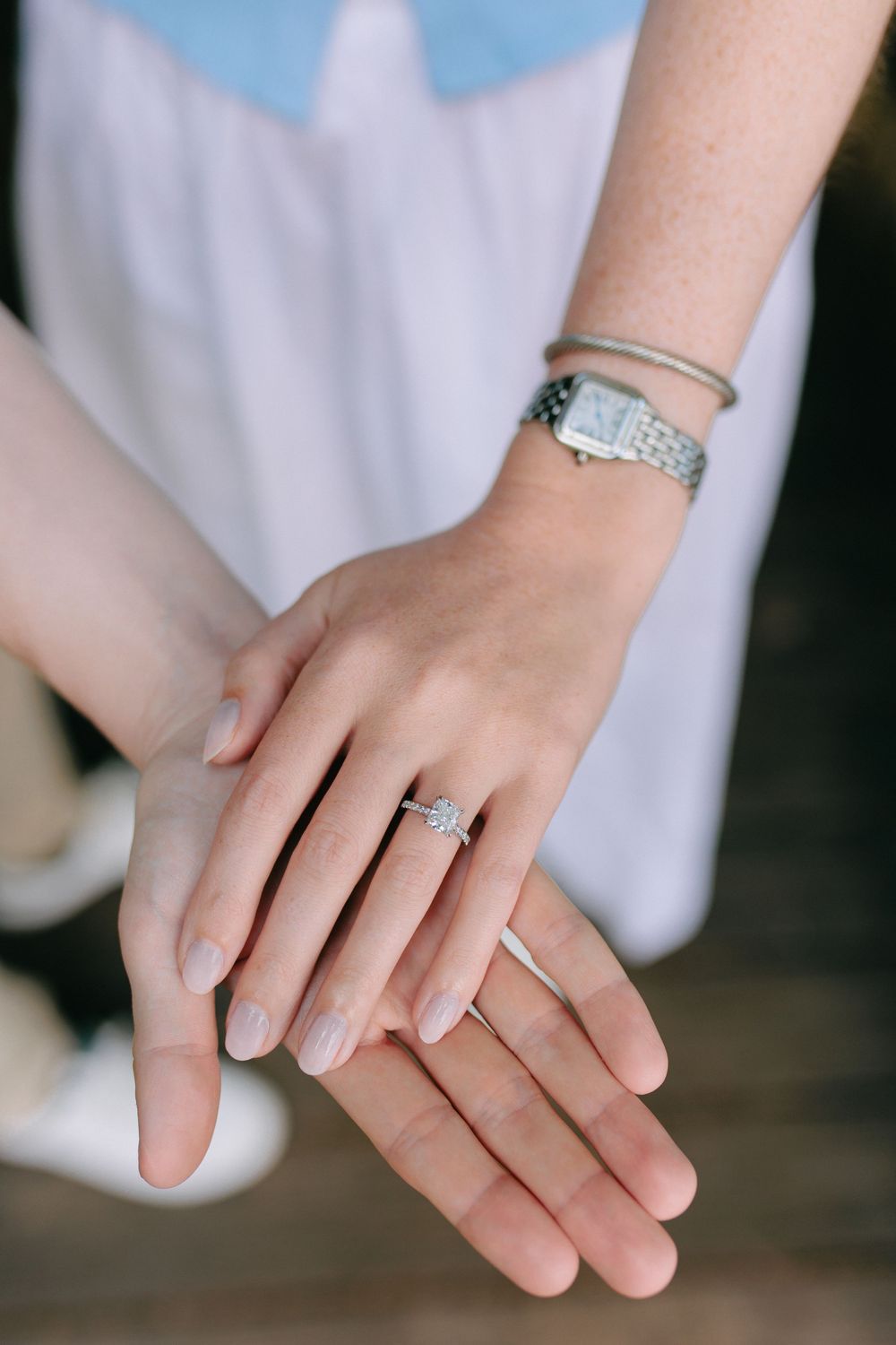 Close up of hands showing an engagement ring and watch against a white dress background.