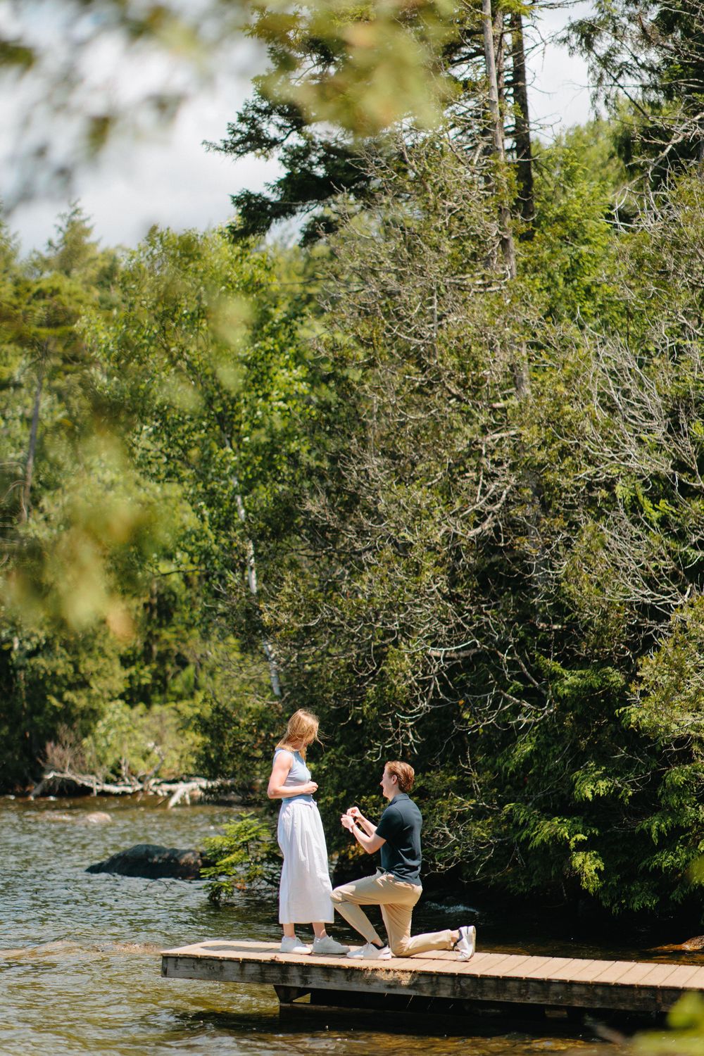 A romantic proposal moment takes place on a wooden dock by a serene lake surrounded by evergreen trees.